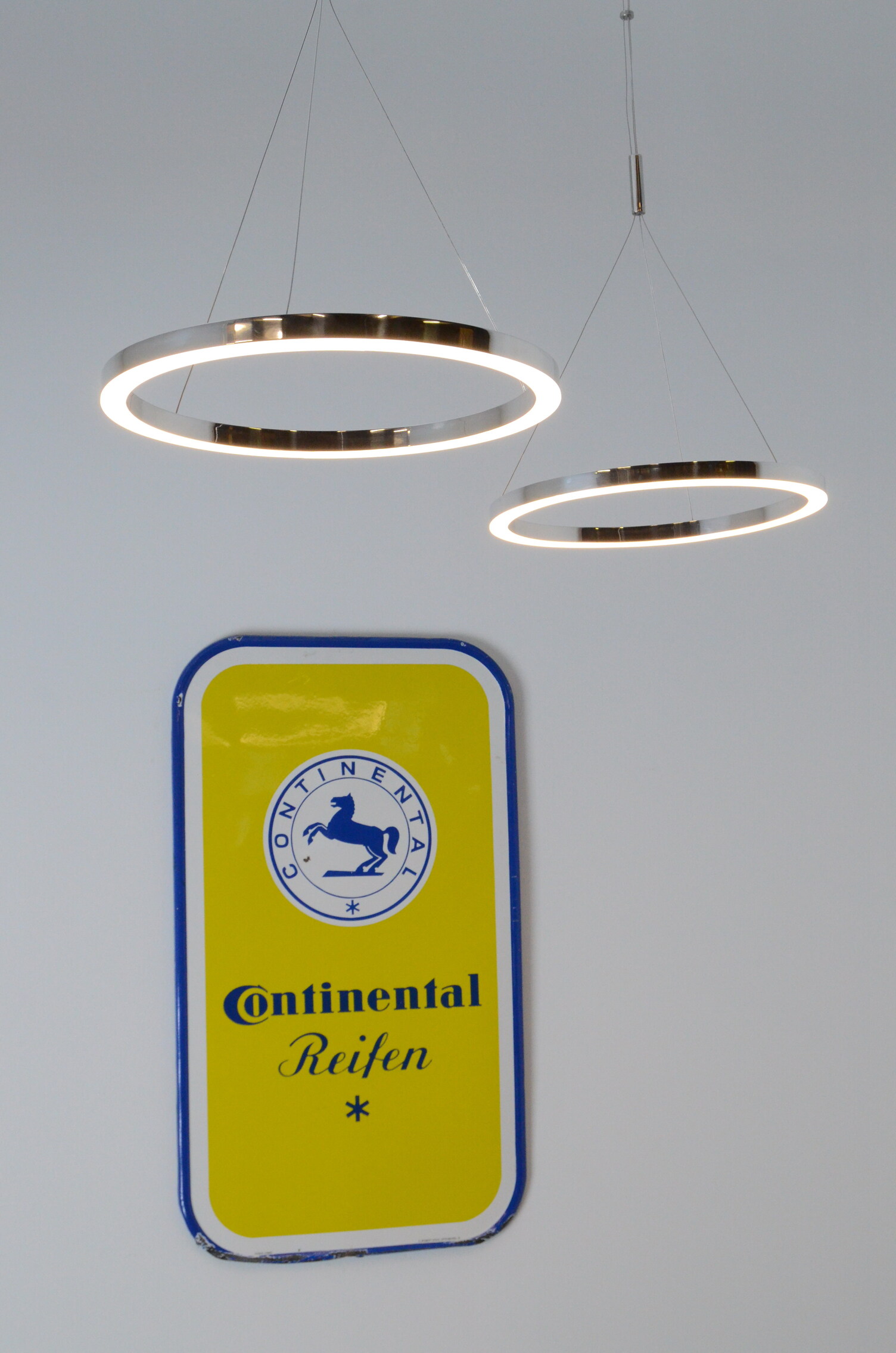 Two small polished aluminium ring lights suspended at different heights in front of a Continentale metal shield. TRIS pendant light.