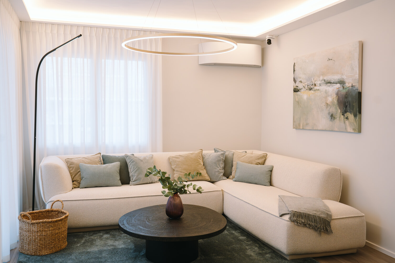 Dezente moderne Wohnzimmerleuchten Modernes Wohnzimmer mit einem blauen Teppichboden. Den Mittelpunkt des Raumes bildet ein helles Sofa mit pastellfarbenen Kissen. Das Zentrum wird markiert durch eine dekorative Wohnzimmerlampe, der Ringleuchte SLIM DIRECT. Weiteres dekoratives und Lichtvolles Element, das über dem Sofa gebogen hervorragt ist die Wohnzimmerstehleuchte PALITO FLOOR, die an eine Peitschenleuchte erinnert. Rechts an der Wand hängt ein schwarz weiß Bild.
