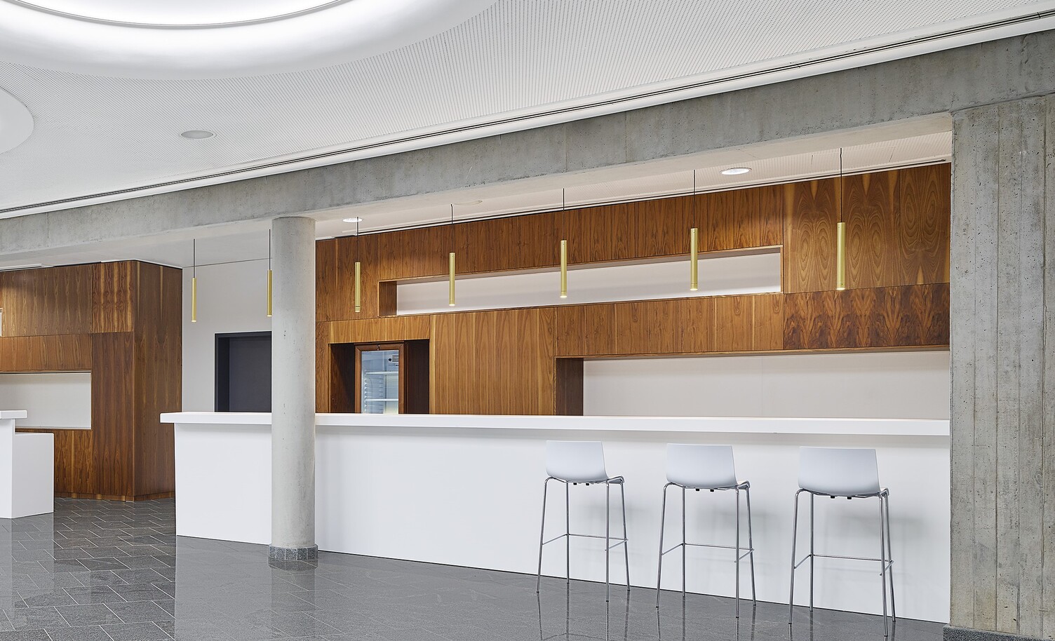 Black Sea Arena Georgia Zoom on counter in a large hall with stone floor and a white ceiling in which large round light fields are arranged, view goes into a counter area, which is characterised as niches and is highlighted by brown wooden walls, in addition, golden bar lights (TIAs) are suspended at regular intervals over the entire counter.