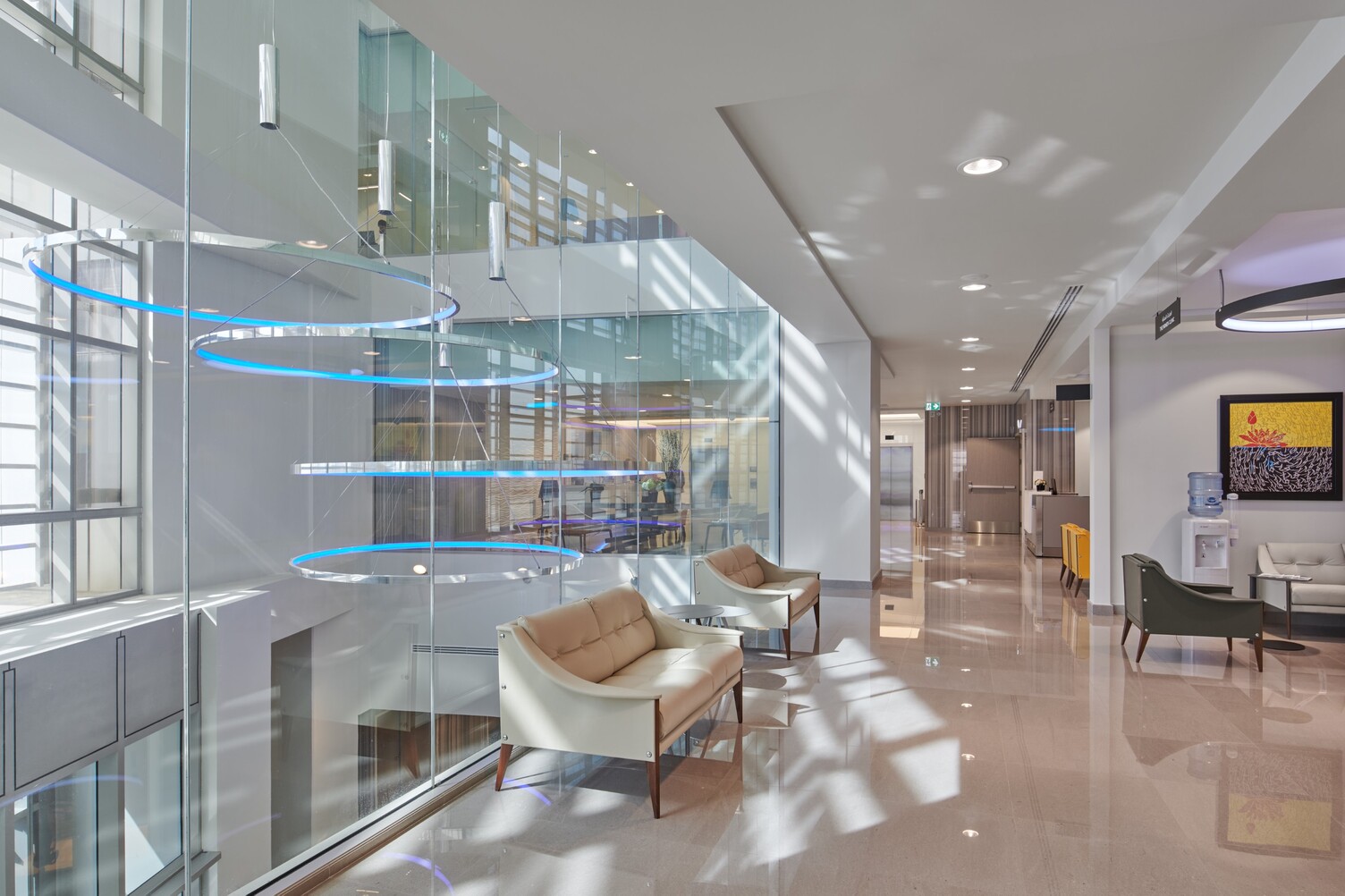 Krankenhausflur Clemenceau Medical Center Dubai mit CIRCOLO Ringleuchten Krankenhausflur, links im Bild Blick auf ein Glasatrium mit vier großen Aluminiumpolierten Ringleuchten CIRCOLO-I mit Pendelbaldachin, die in blau leuchten und in unterschiedlichen Höhen abgependelt sind.