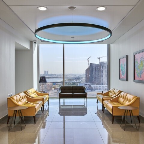 View into a waiting area with a large window front and a view of Dubai's skyscrapers; a round black pendant ring light can be seen shining blue above the furniture in the centre of the area.
