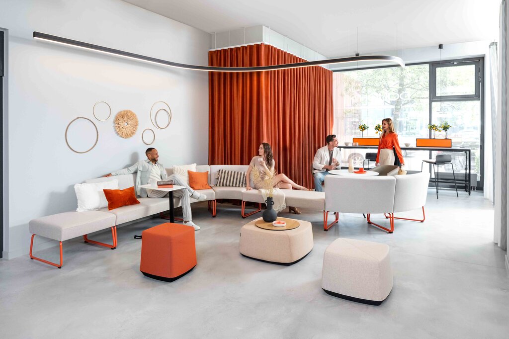 Stylish lounge area in a modern showroom with orange accents and Sattler's Duetto curved lighting fixture in Offenbach. | ©  Dauphin HumanDesign® Group GmbH & Co. KG