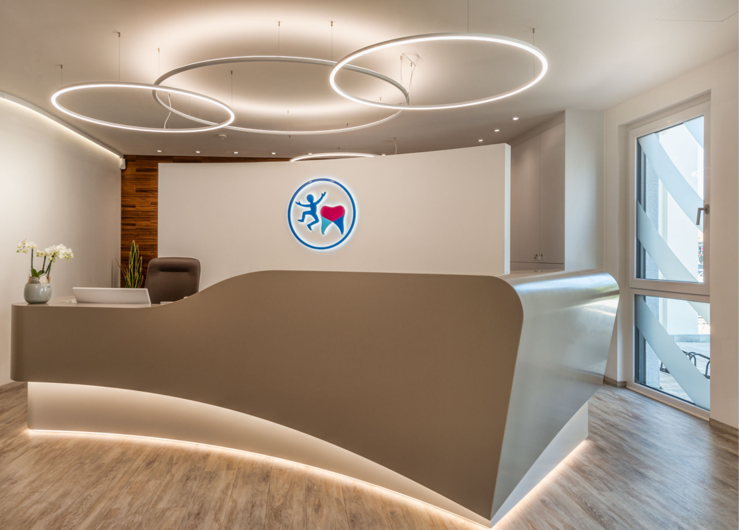 SLIM Ringleuchten über Empfangstresen Beige geschwungene Empfangstheke Kieferorthopädiepraxis Dr. Grammatidis, bodennah abgesetzt und hinterleuchtet, auf der Rückwand ist ein Zahn und ein Strichmännchen, über der Theke hängen 3 schmale SLIM Ringleuchten von SATTLER versetzt angeordnet, zwei leuchten direkt, eine leuchtet indirekt, und hellt die Decke auf.
