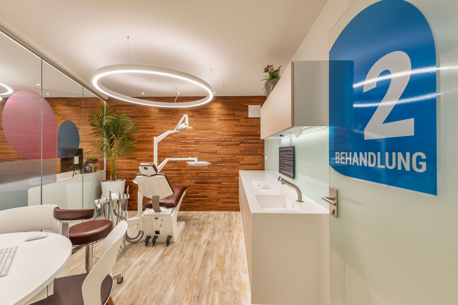 Ringleuchte DUETTO MED über einem Zahnarztbehandlungsstuhl Moderner warmes Behandlungszimmer bei Dr. Grammatidis, Kirchheim; Fachzahnärzte für Kieferorthopädie, mit Fokus auf einen Zahnarztbehandlungsstuhl über dme eine weiße Ringleuchte DUETTO MED abgependelt ist.