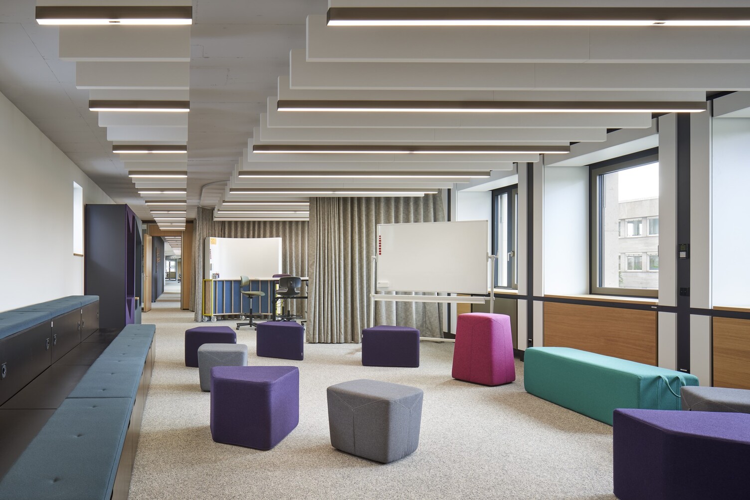 New Work workshop area with baffle luminaires Workshop office space with colourful upholstered furniture and seating cubes, above which several linear acoustic baffles and lights can be seen in two rows one behind the other.