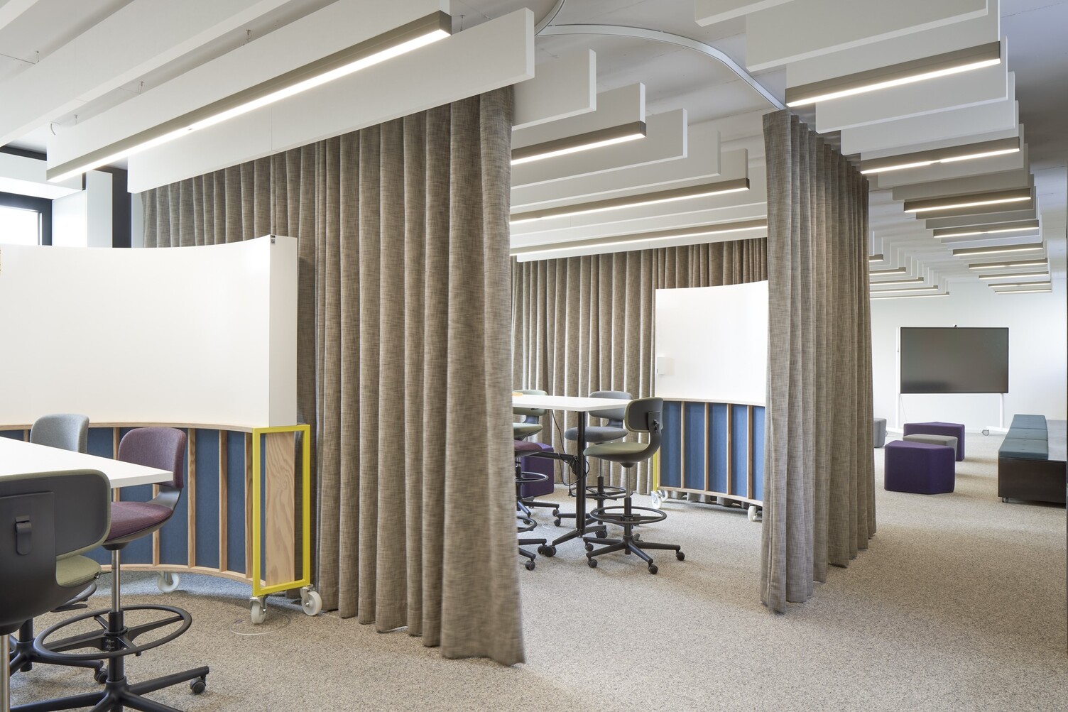 New Work workshop area with room units that can be separated by curtains; linear acoustic baffles are arranged in rows on the ceiling throughout.