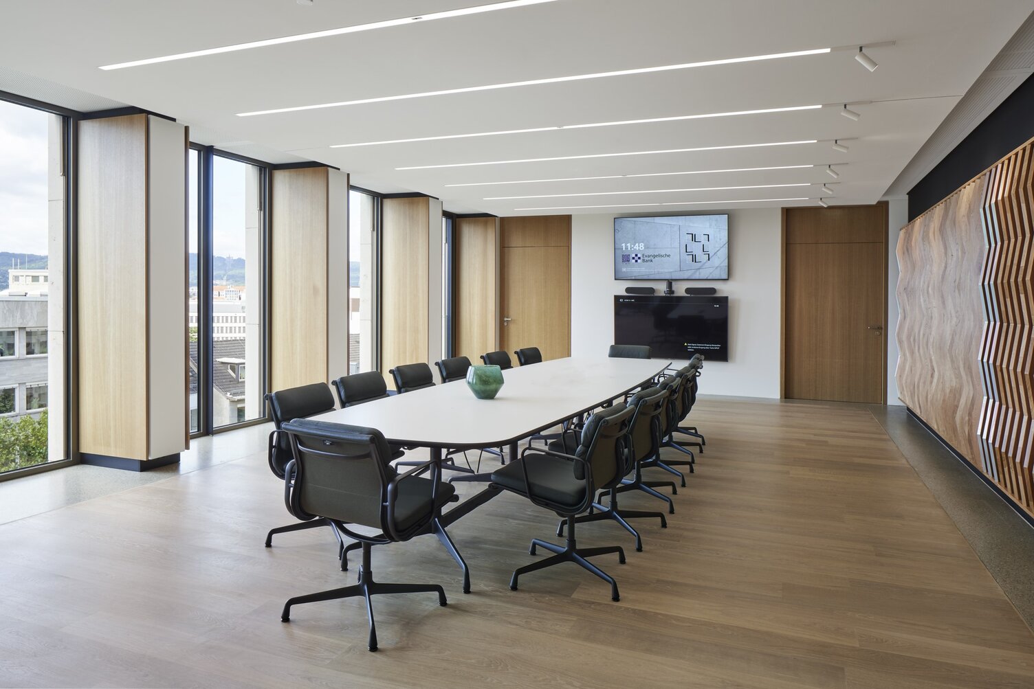 Konferenzraum der EVB mit einem ovalen Meetingtisch und Bürostühlen darum herum in der Mitte, außenherum geben Fenster den Blick nach draußen frei, in der weißen Decke sind lineare Einbauleuchten (PALADIO) sichtbar, zu einer Wandseite sind am Ende der Leuchten noch zylindrische Spots (EVOLUTION SPOT) anschließend eingebaut.