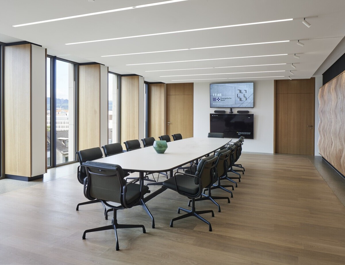 EVB conference room with an oval meeting table and office chairs around it in the centre, windows around the outside provide a view of the outside, linear recessed luminaires (PALADIO) are visible in the white ceiling, and cylindrical spotlights (EVOLUTION SPOT) are installed at the end of the luminaires on one side of the wall.