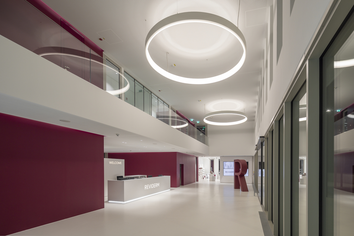 Eingangsbereich Headquarter Reviderm München in Rottönen und weiß gehaltene Wände links im Bild, gegenüberliegend Fensterfront, mittig im Gang mit großen CIRCOLO Ringleuchten in weiß nach innen leuchtend.