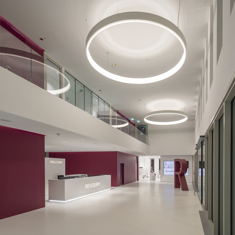Eingangsbereich Headquarter Reviderm München in Rottönen und weiß gehaltene Wände links im Bild, gegenüberliegend Fensterfront, mittig im Gang mit großen CIRCOLO Ringleuchten in weiß nach innen leuchtend.
