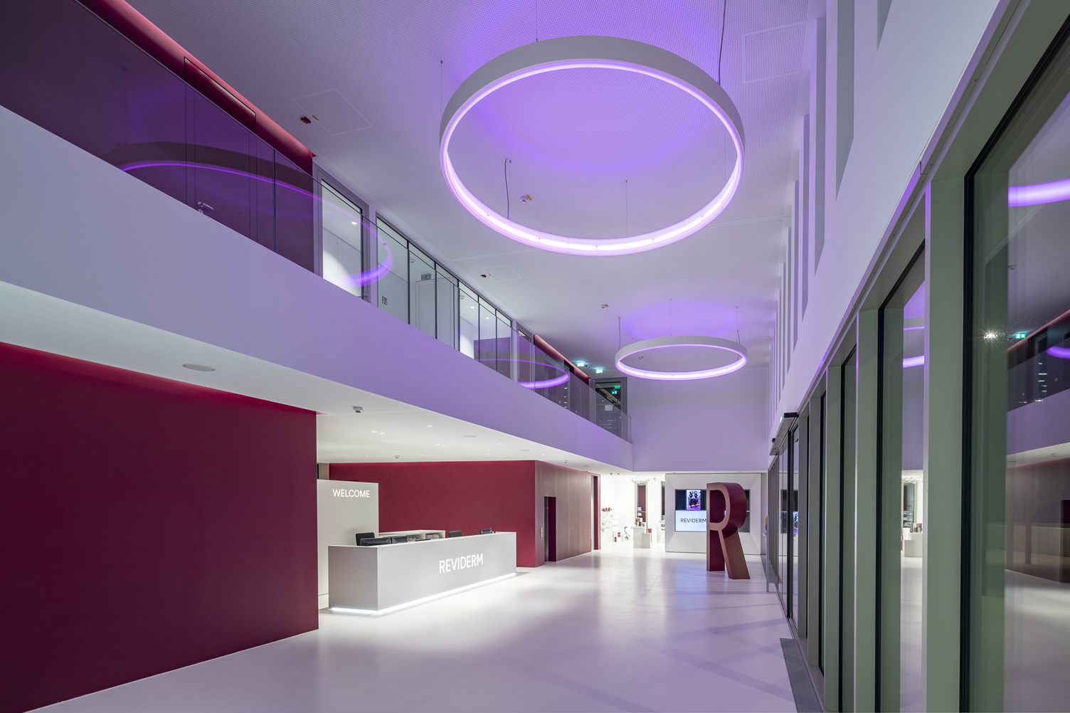 Ringleuchte CIRCOLO mit RGB im Eingangsbereich Reviderm Foyer in Unternehmensfarben von Reviderm in weiß und und einem warmen Rotton. Entlang des Flurs an der Empfangstheke vorbei hängen CIRCOLO Ringleuchten, die in einem lila Ton leuchten.