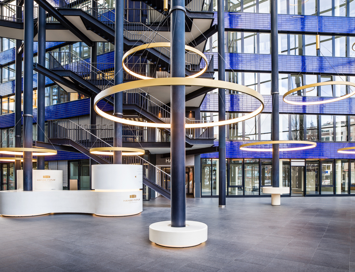 CIRCOLO Ringleuchten um Säulen des Atriums Hanseforum Hamburg Goldene großformatige Ringleuchten, CIRCOLO I, abgependelt um die Säulen des verglasten Atriums mit blauen Wänden des Hanseforum Hamburg.