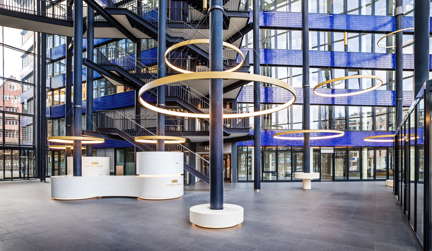 CIRCOLO Ringleuchten um Säulen des Atriums Hanseforum Hamburg Goldene großformatige Ringleuchten, CIRCOLO I, abgependelt um die Säulen des verglasten Atriums mit blauen Wänden des Hanseforum Hamburg.