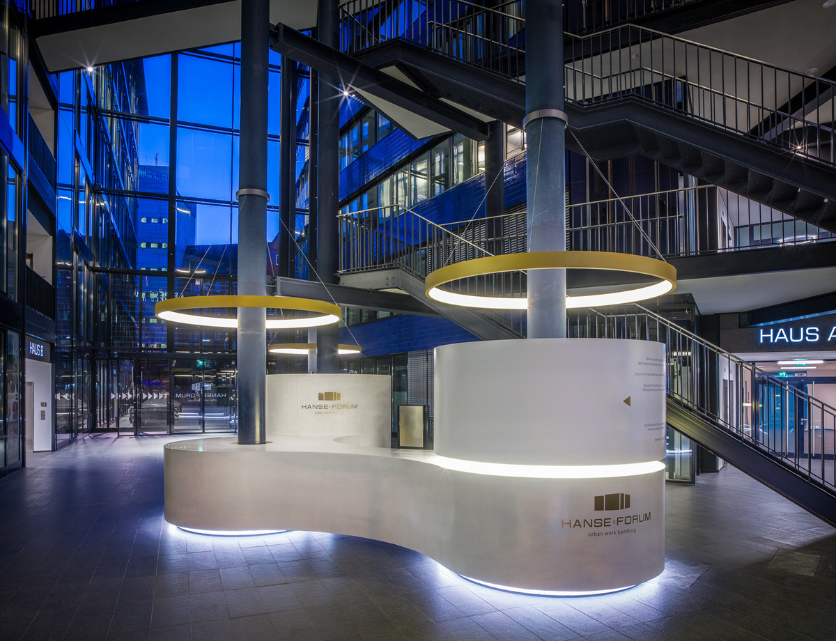 Goldene Ringleuchten abgependelt an Säulen über Empfangstheke Verbundene s-förmige, weiße, runde Empfangstheke von stayconcrete im Atrium des Hanseforum Hamburg um zwei blaue Säulen, von diesen Säulen ist je eine goldene Ringleuchte CIRCOLO schirmartig abgependelt.