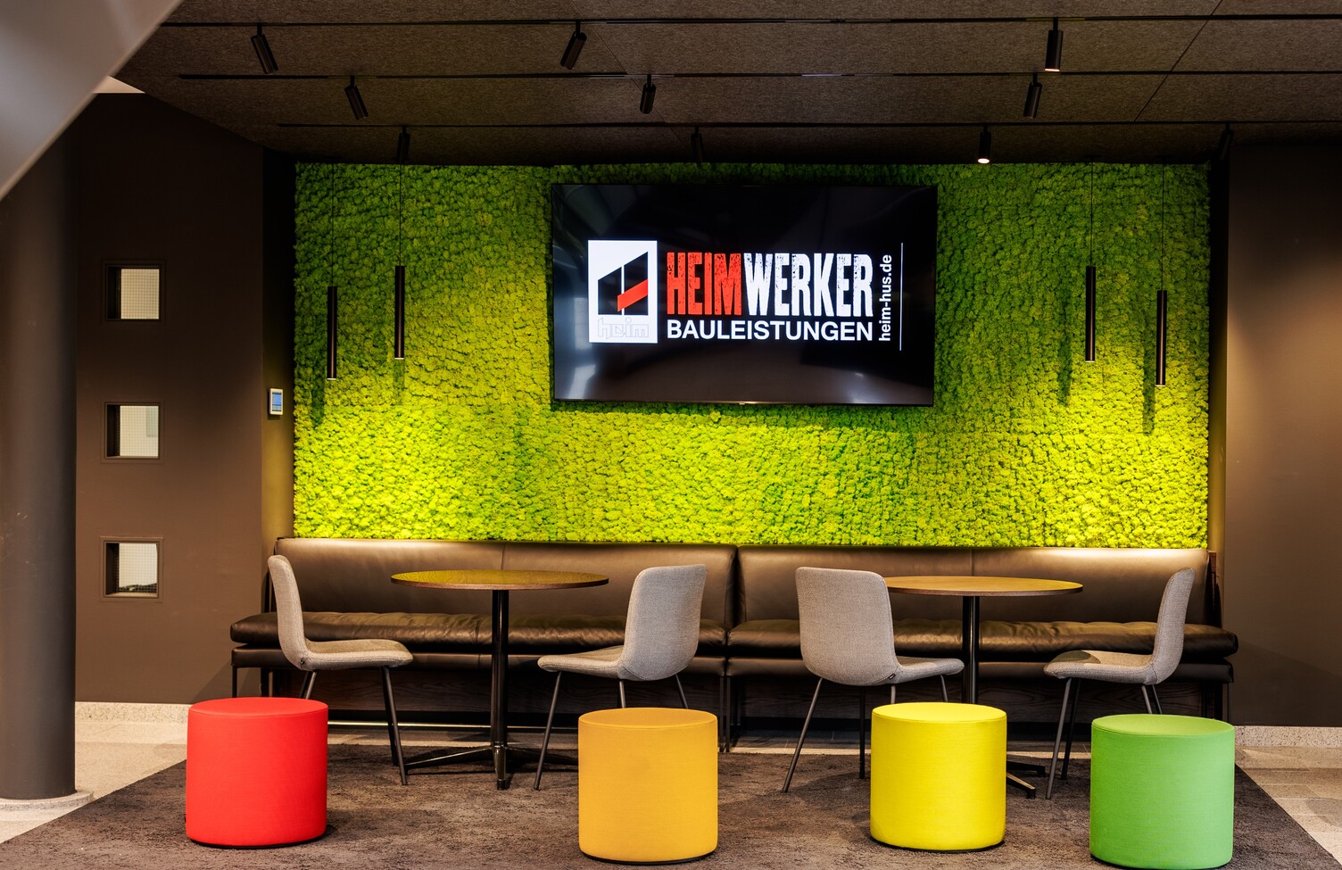 Lounge Heim Infrastrukturbau | Gaus Architekten Colourful waiting lounge at Heim Infrastrukturbau with a green wall in front of which cylindrical pendant luminaires (TIA) are suspended and a light track system with spotlights on the ceiling.