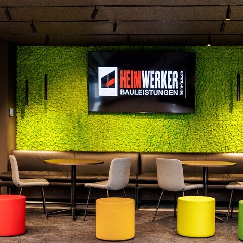 Lounge Heim Infrastrukturbau | Gaus Architekten Colourful waiting lounge at Heim Infrastrukturbau with a green wall in front of which cylindrical pendant luminaires (TIA) are suspended and a light track system with spotlights on the ceiling.