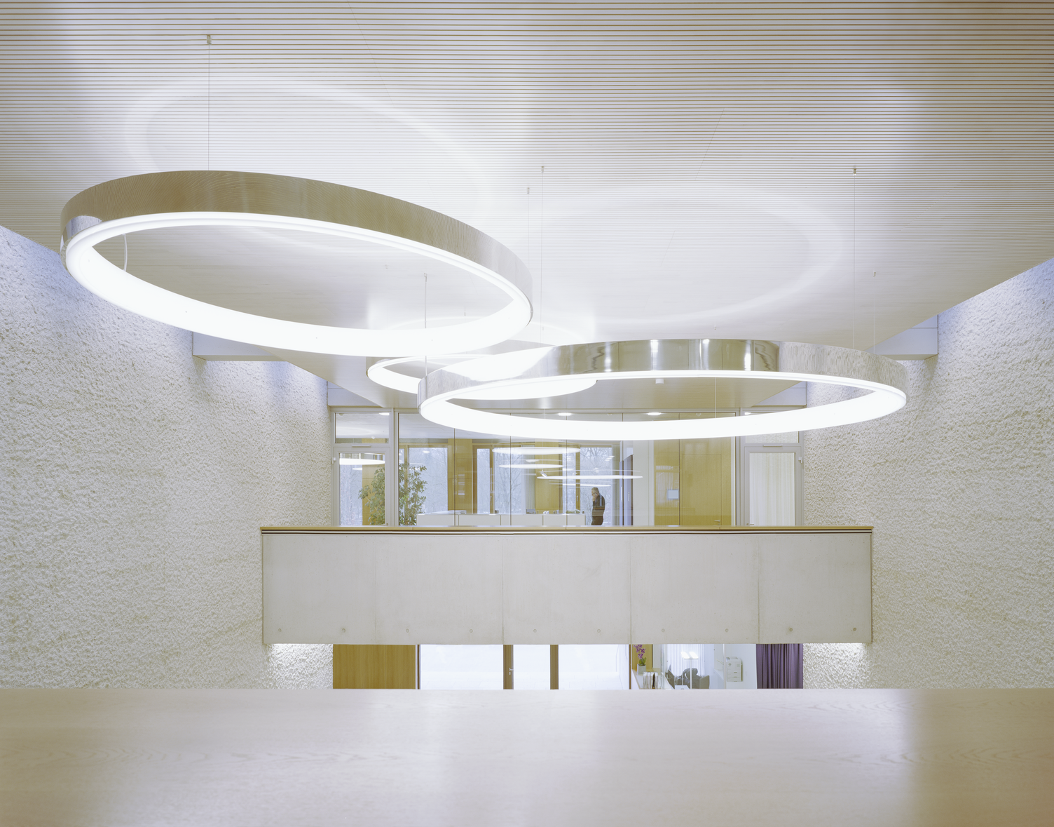 Ring lights in the exposed concrete building of Karl Köhler GmbH Three large-format CIRCOLO ring luminaires in polished aluminium as a focal point in the exposed concrete building combined with light-coloured wooden elements from Karl Köhler GmbH. Suspended close to the ceiling at 1st floor level of the glazed office. Free space under the luminaires can be used as an event room.