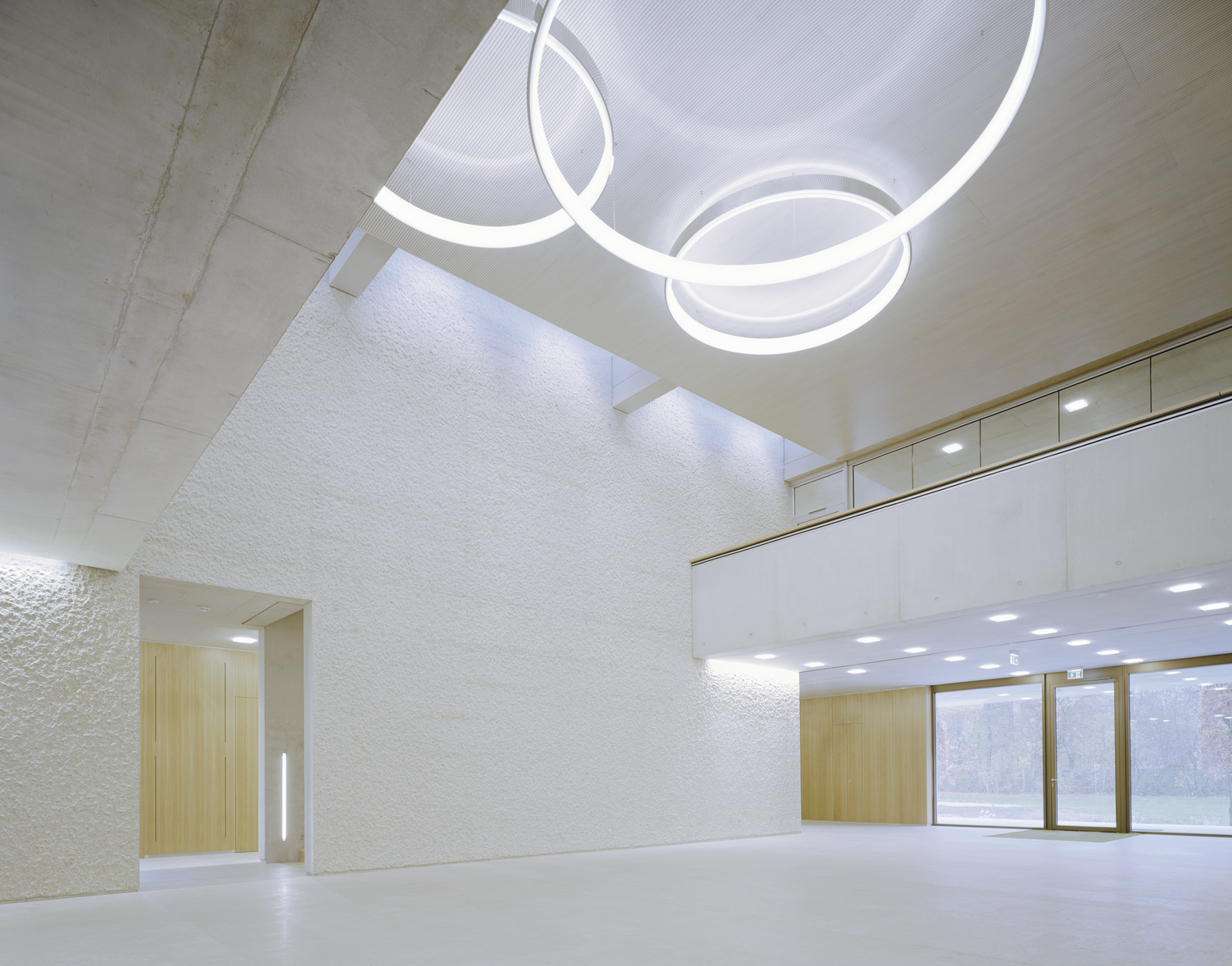 Venue & meeting point under CIRCOLO ring lights View from below to the ceiling of an exposed concrete building, 3 CIRCOLO ring luminaires in polished aluminium hang in the middle of the two-storey high room, on both sides the room narrows by one storey.