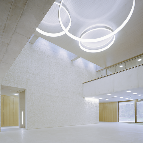 Venue & meeting point under CIRCOLO ring lights View from below to the ceiling of an exposed concrete building, 3 CIRCOLO ring luminaires in polished aluminium hang in the middle of the two-storey high room, on both sides the room narrows by one storey.