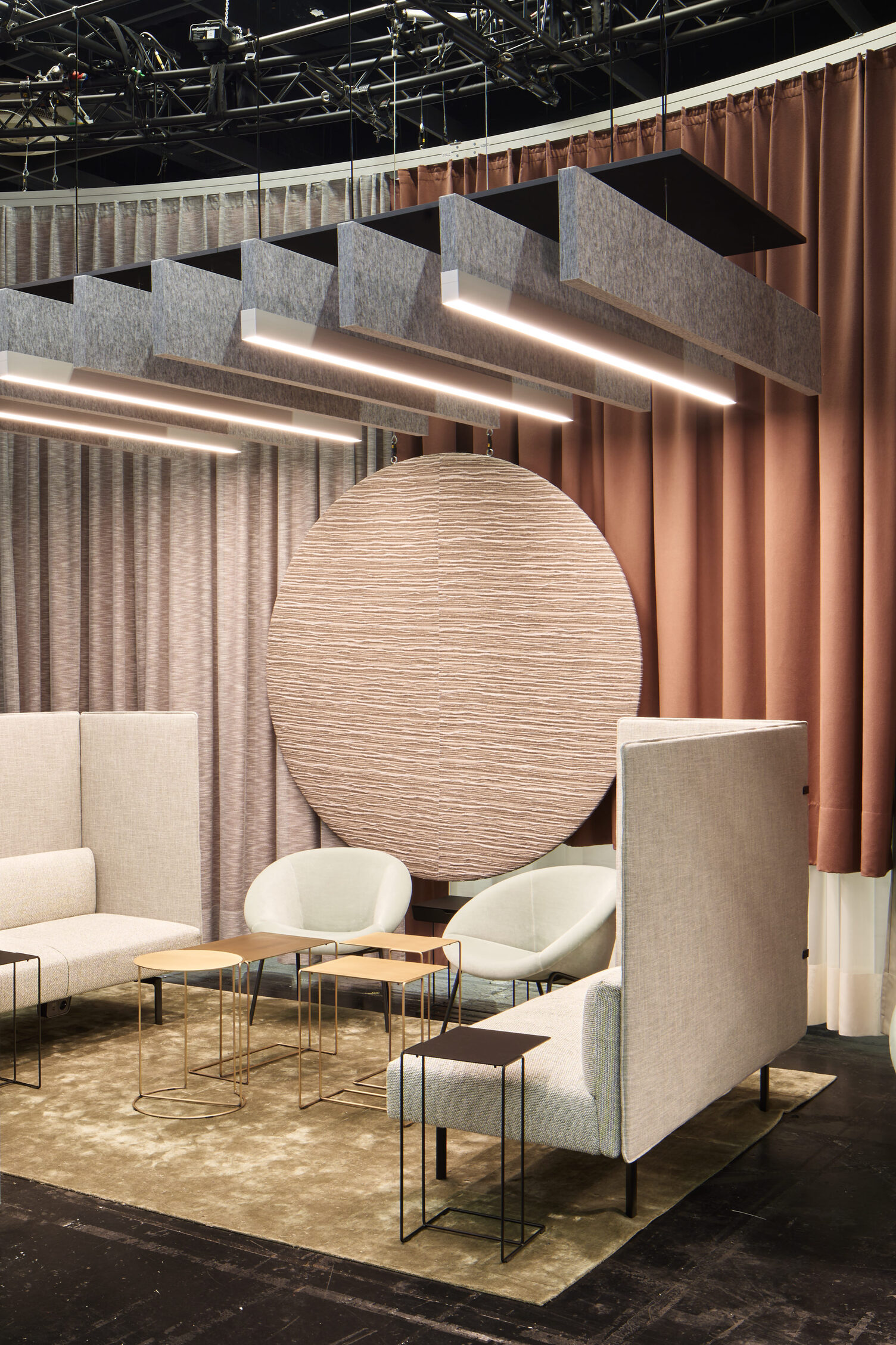 Acoustically effective room-in-room solution with BAFFELLEUCHTEN ORGATEC 2024 exhibition stand by Walter Knoll with light-coloured upholstered furniture and small sofa tables, above which several linear baffle luminaires (BAFFELLEUCHTE) and pure linear acoustically effective baffles (ACOUSTIC BAFFLE) in grey are suspended.