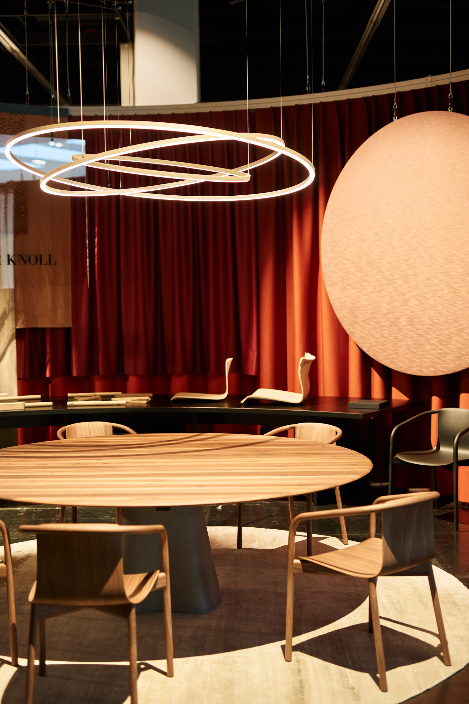 PURE MOTION SLIM über rundem Tisch bei WALTER KNOLL Messestand von WALTER KNOLL auf der ORGATEC mit einem runden Holztisch und Holzstühlen darum, darüber schweben drei ineinander verkreuzte Ringleuchten, die PURE MOTION.