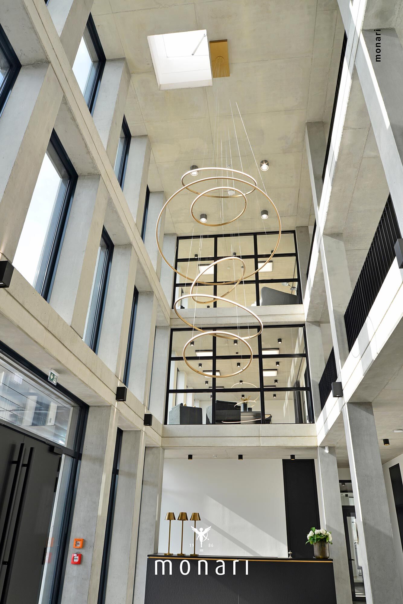 Stilvoller Kronleuchter über der Empfangstheke von Monari Moderner Eingangsbereich eines architektonisch anspruchvollen Betongebäudes von Monari. Geschmückt durch schwarze Fensterrahmen. Blick auf die Empfangstheke mit insbesonderem Augenmerk auf den individuellen goldenen Kronleuchter aus 6 SLIM Ringleuchten nach außenstrahlend über der Empfangstheke.