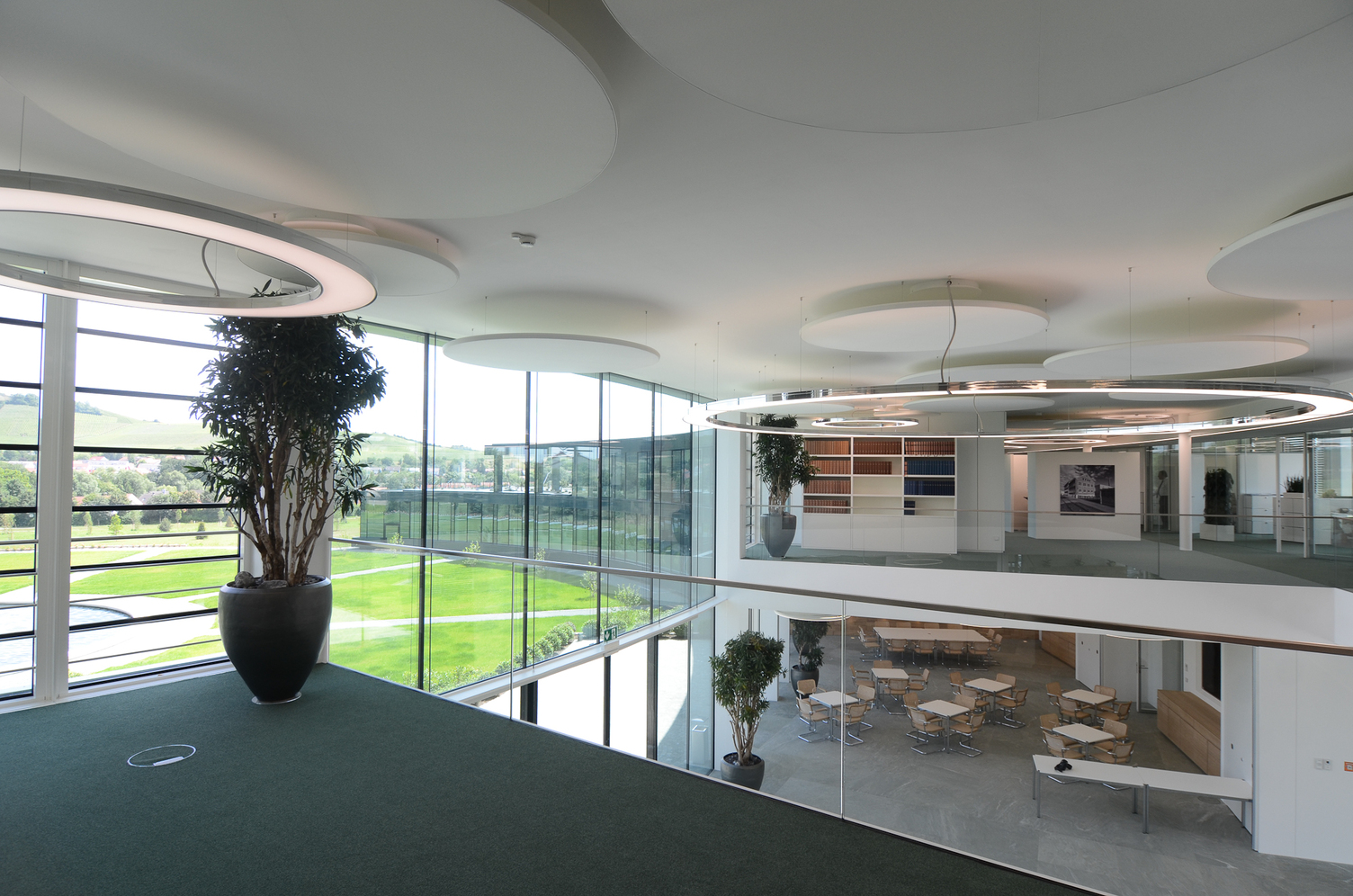 Anello und Acoustic Lights von SATTLER in Kombination in einem Foyer Blick von einer Empore in ein modernes Foyer mit Glasfront. Ausgestattet wurde das Foyer mit Anello Leuchten, stoffbespannte Akustik Panele, jeweils von SATTLER.