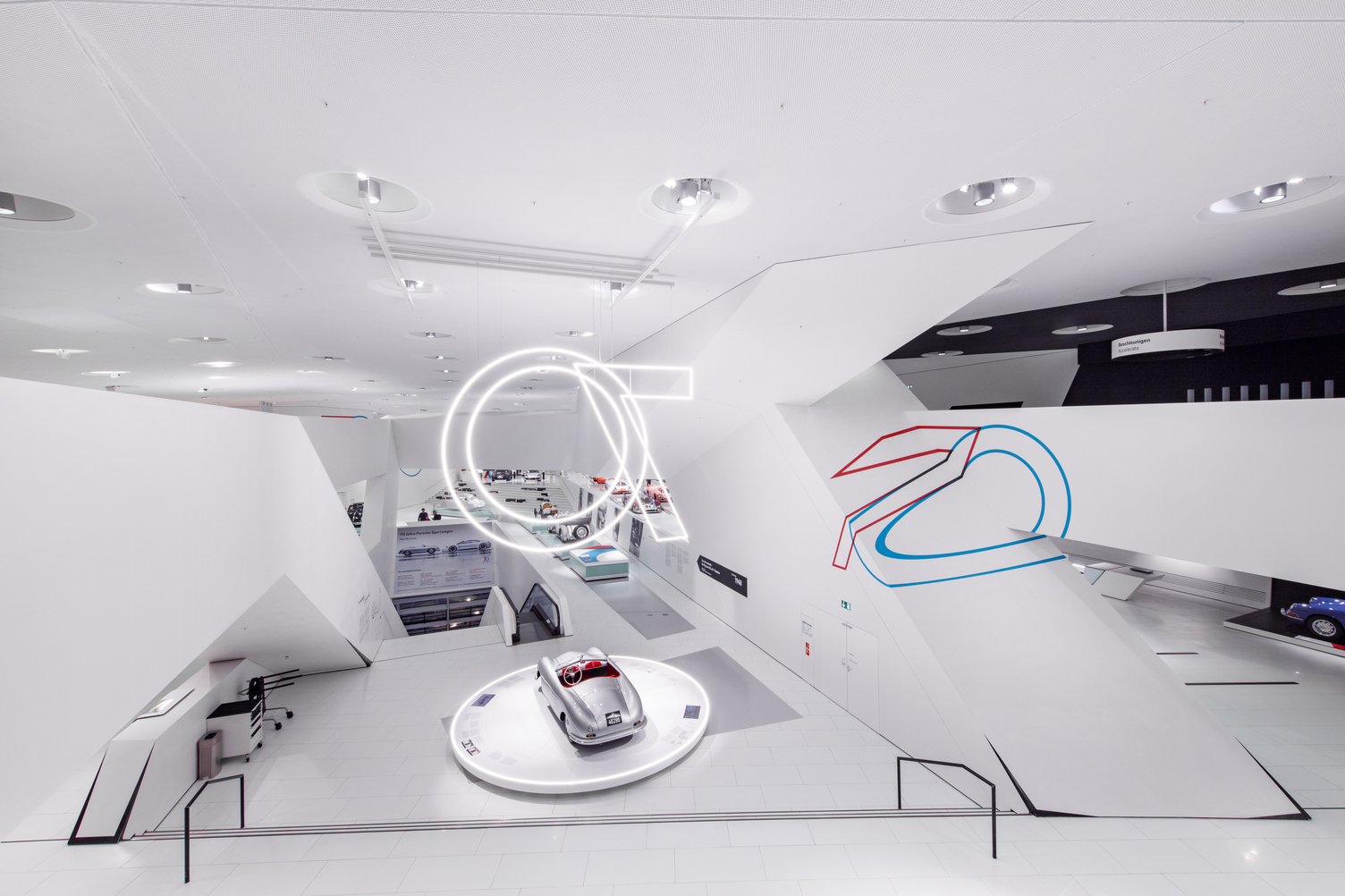 Wide interior view of the Porsche Museum to mark 70 years of Porsche sports cars in Stuttgart, showing the exhibition space with modern lighting from Sattler. For this purpose, a 70 was formed from the Doppio luminaire series from SATTLER and displayed from the ceiling above the sports cars. Doppio Linea and Doppio round luminaires were used, each suspended vertically from the ceiling to be visible from all sides of the building inside.
