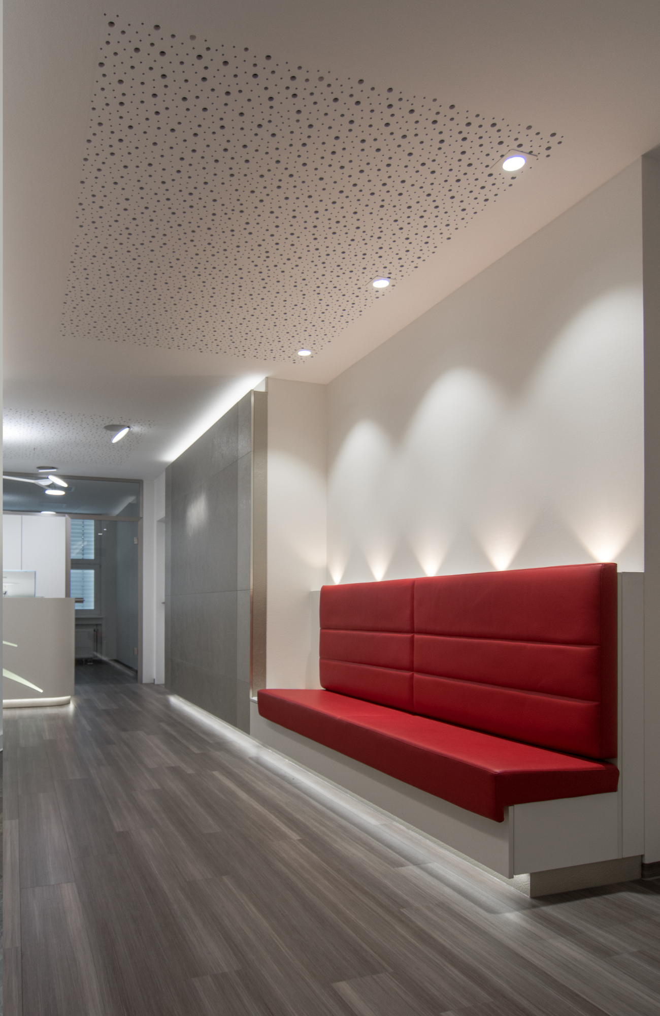 Praxis Wartebereich im Flur mit einer roten Bank, hinter der fünf lichtkegel nach oben strahlen, und drei Spots an der Decke nach unten auf dei Bank, im Hintergrund ist der Empfangs-Tresen zu sehen.