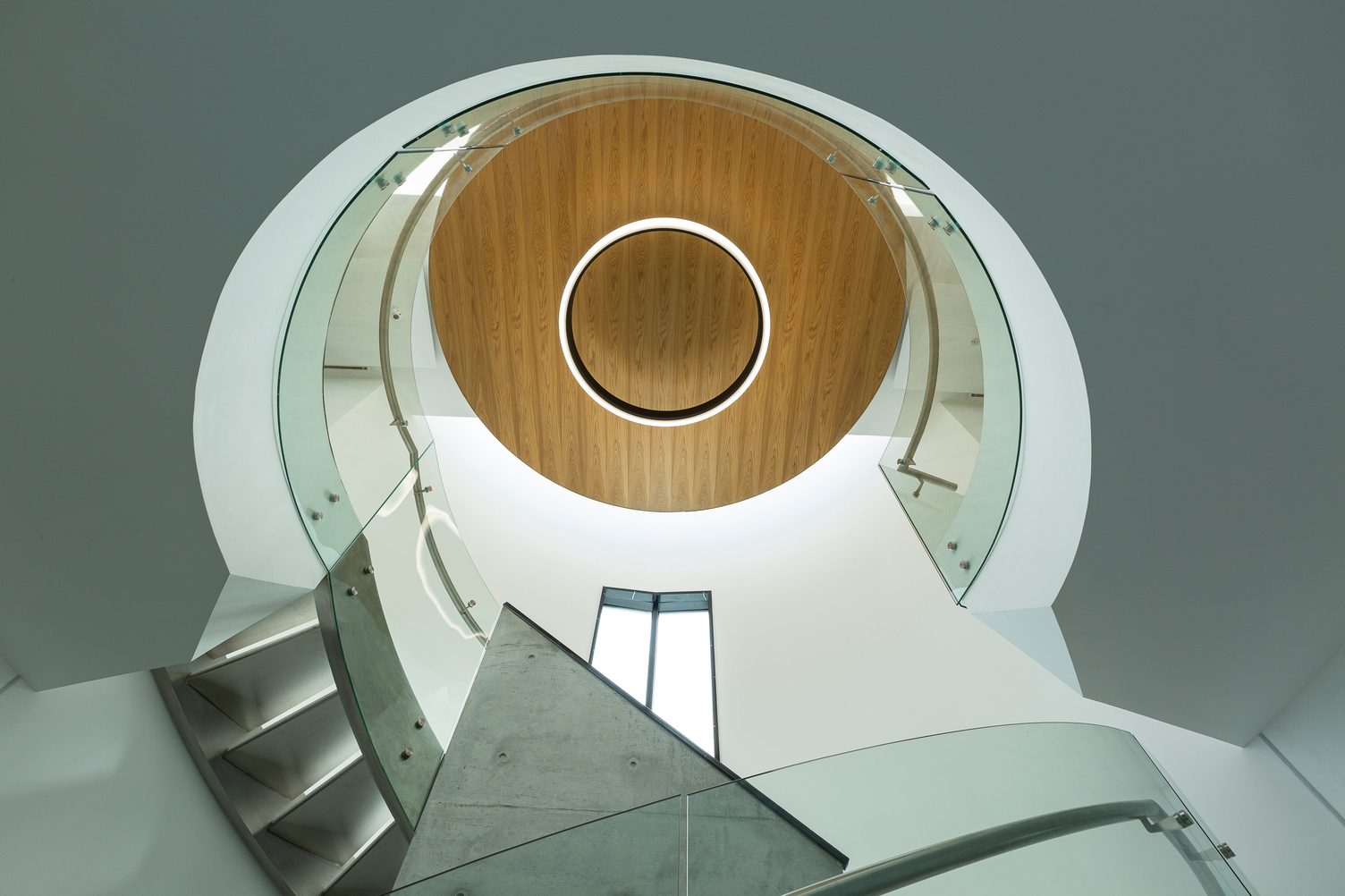 Beleuchtung des Treppenauges mit großformatiger Ringleuchte Blick nach oben in einem Treppenhaus mit Glashandlauf und im Mittelpunkt an Holzdecke eine runde nach außen Strahlende Leuchte, CIRCOLO INVERSE Dmr 1600.