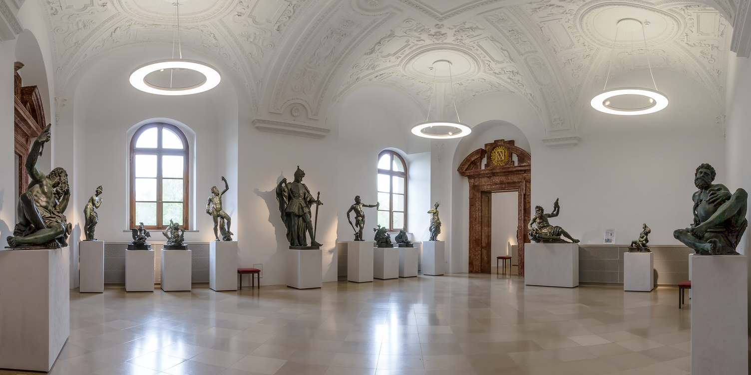 ANELLO Ringleuchten mit Spots in den Bronzesälen der Residenz München Bronzesaal der Bayerischen Schlösserverwaltung in der Residenz München, Ausstellungsraum mit Bronze-Skulpturen auf weißen Podesten, von der Stuckdecke mit Gewölbestruktur sind Ringpendelleuchte ANELLO mit integrierten Spots abgependelt, die die Ausstellungsstücke und den Raum ausleuchten.