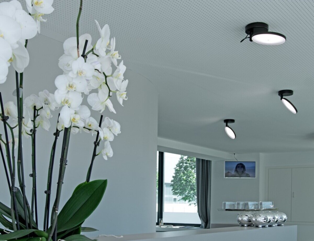 Bewegliche Deckenanbauleuchten für Empfangstheken Weiße geteilte lineare Empfangstheke im Friseursalon Mentes. Links steht eine weiße Orchidee auf dem Tresen, an der weißen Lochdecke darüber sind entlang der Thekenlänge einzelne schwarze AVVENI SINGLE, kleine Anbauleuchten mit beweglichen runden flachen Leuchtenköpfen, die in verschiedene Richtungen ausgerichtet sind.