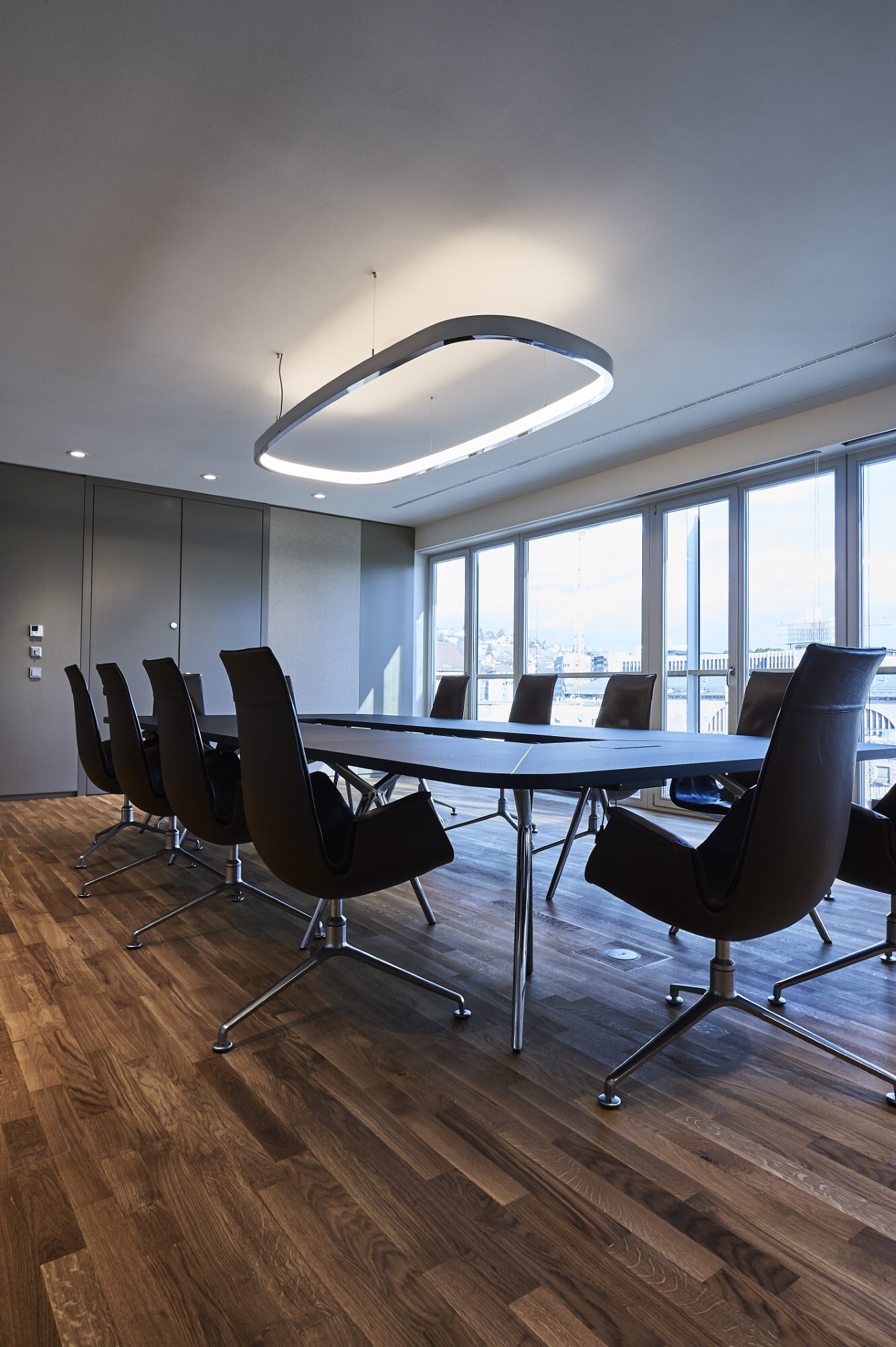 Besprechungsraum Beleuchtung Blick von einem Eck eines rechteckigen Besprechungstisches schräg nach oben an die Decke, von der eine Ringleuchte in Form eines abgerundeten Rechtecks abgependelt ist. Außen in poliertem Aluminium innen mit einem Diffusor, leuchtet sie nach innen und dabei erhellt sie sowohl die Decke, als auch den Tisch.