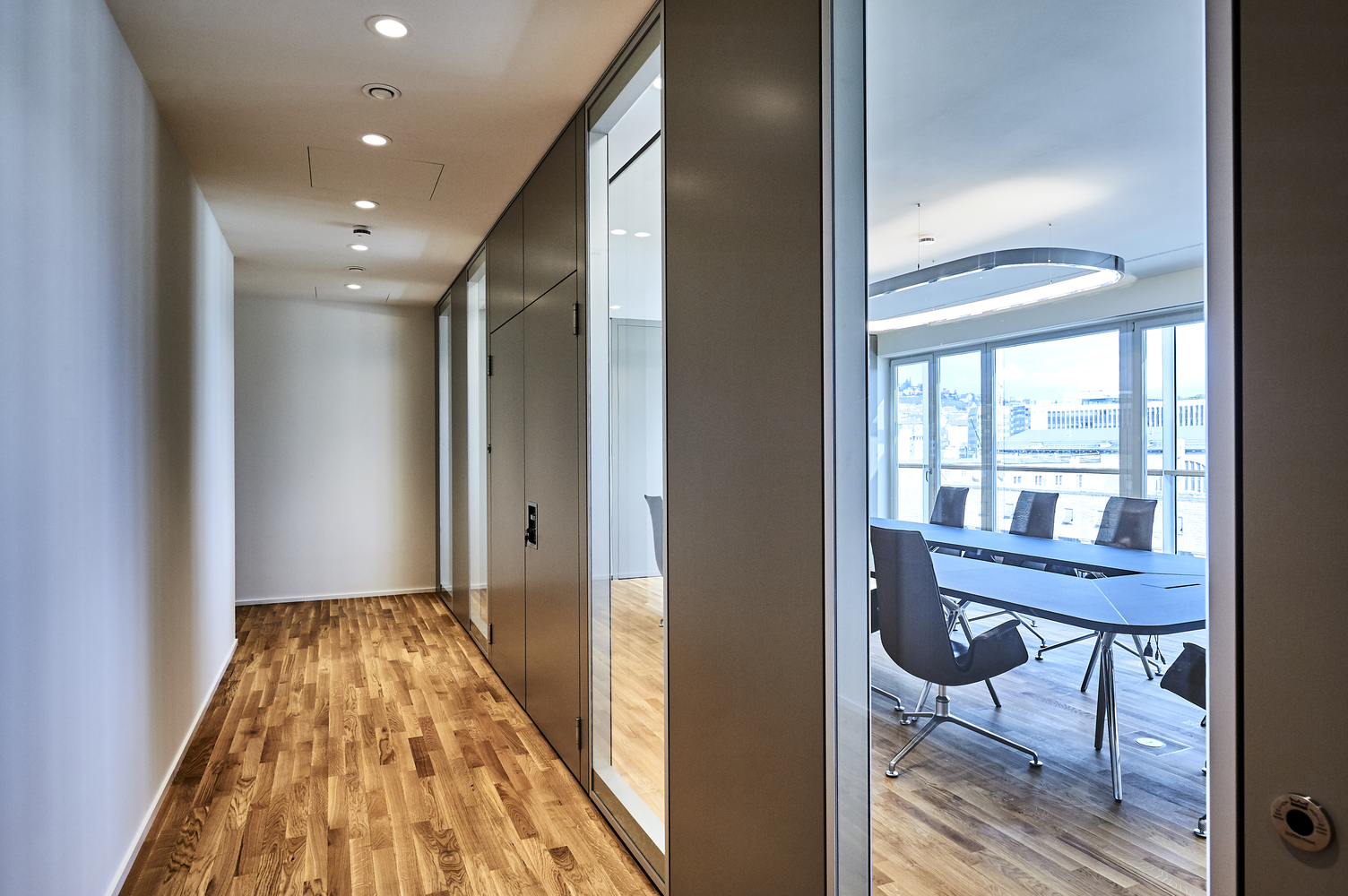 Corridor lighting Corridor in a modern office with strip parquet flooring, illuminated by small round recessed spotlights, BE2 luminaires. Through a glass front, you can see into an adjacent meeting room with a table, chairs and rectangular SOFT SQUARE ring luminaire.