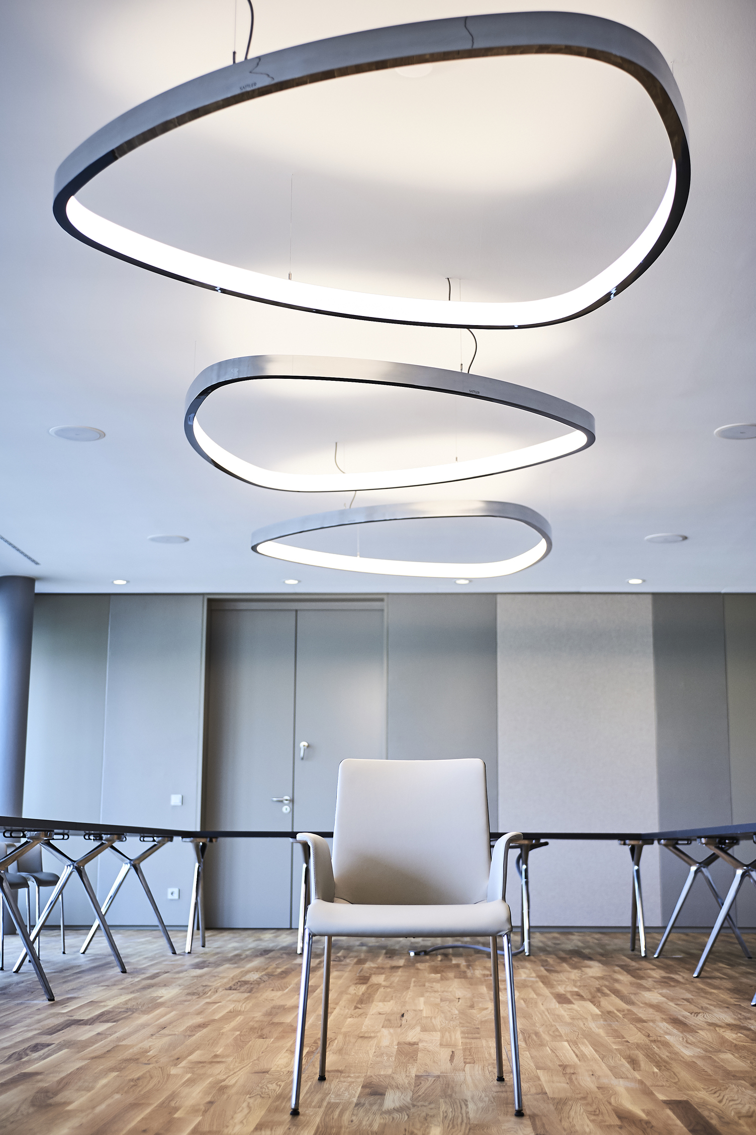 3 dreieckige SOFT DELTA beleuchten WALTER KNOLL Stuhl Drei Aluminium polierte Ringleuchten in Form eines abgerundeten Dreiecks (SOFT DELTA) sind von der Decke in Reihe hintereinander mit verschiedenen Ausrichtungen abgependelt, darunter steht ein heller Stuhl von WALTER KNOLL auf einem braunen Stäbchenparkett.