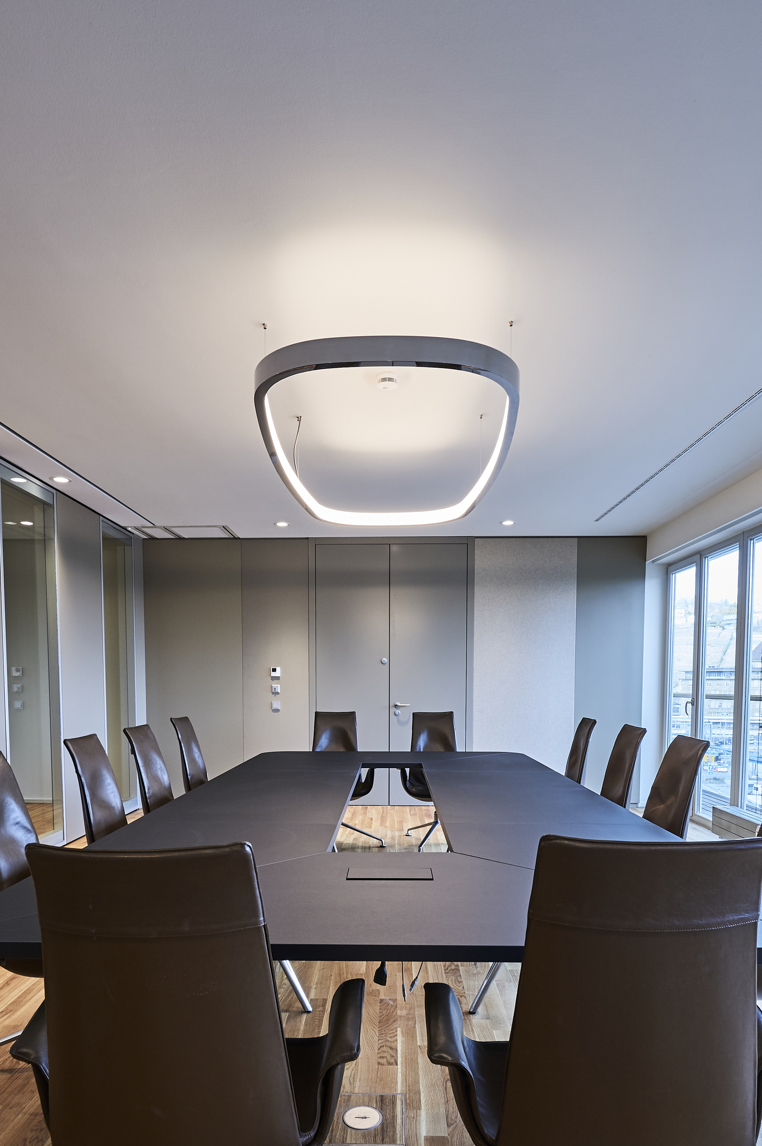 Lighting for rectangular meeting tables View from the short side of a rectangular meeting table diagonally upwards to the ceiling, from which a ring light in the shape of a rounded rectangle is suspended. Polished aluminium on the outside and with a diffuser on the inside, it shines inwards, illuminating both the ceiling and the table.