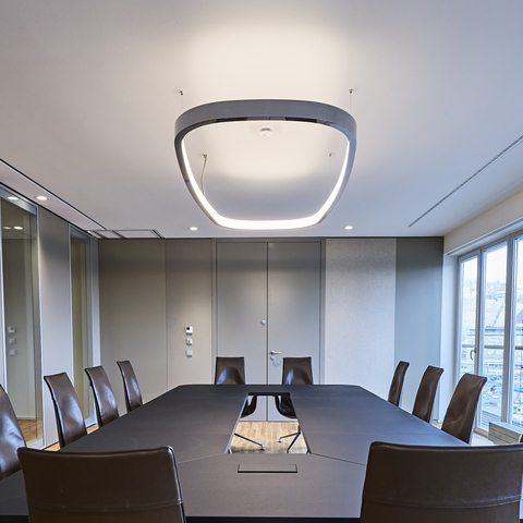Lighting for rectangular meeting tables View from the short side of a rectangular meeting table diagonally upwards to the ceiling, from which a ring light in the shape of a rounded rectangle is suspended. Polished aluminium on the outside and with a diffuser on the inside, it shines inwards, illuminating both the ceiling and the table.