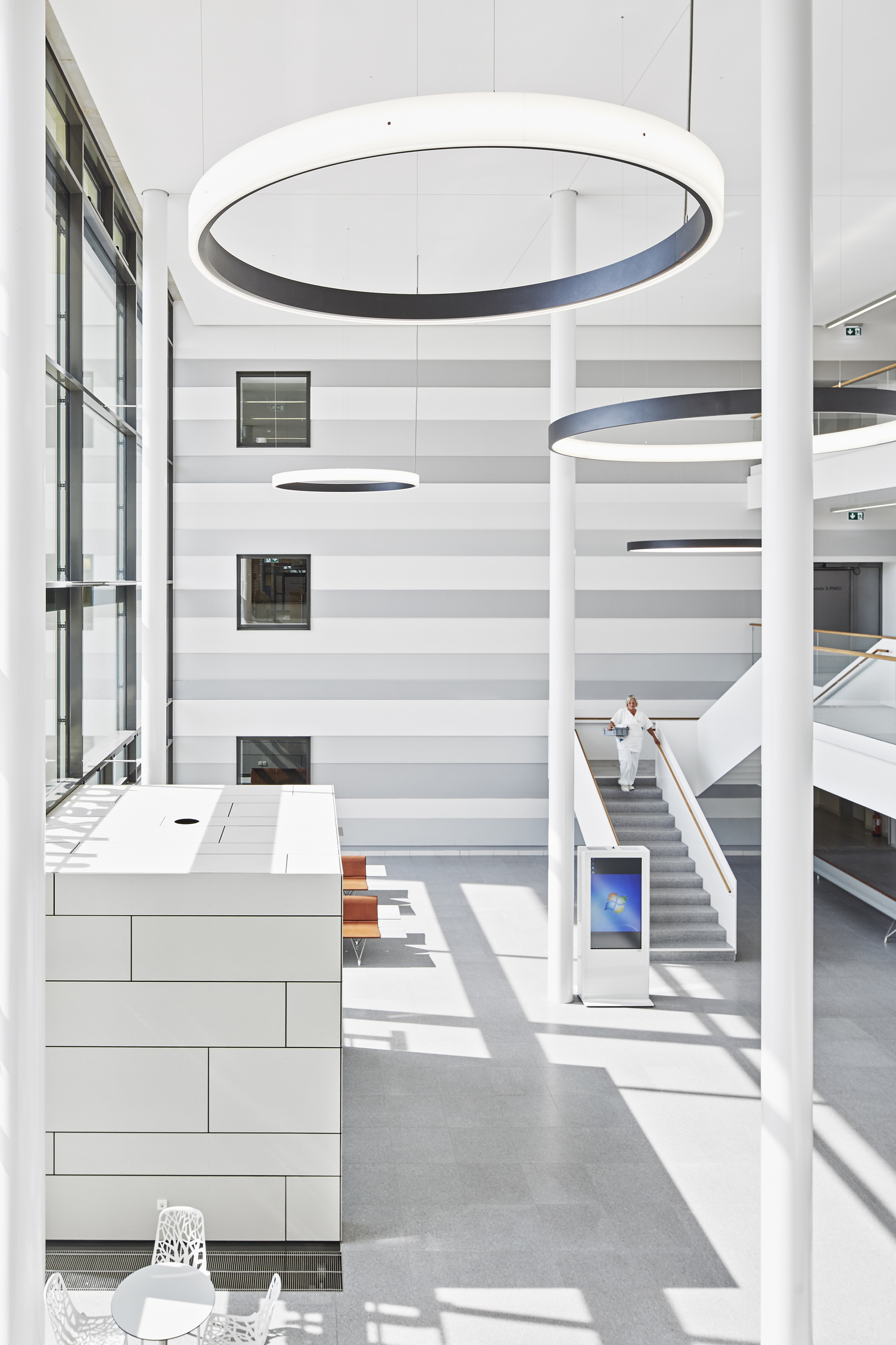 Eingangshalle & Treppenhaus Thoraxklinik mit CIRCOLO Ringleuchten Hohe weiße Eingangshalle der Thoraxklinik mit Säulen, 2 große schwarze Ringleuchten nach innenstrahlend (CIRCOLO I) und 2 Ringleuchten (CIRCOLO INVERSE) nach außenstrahlend im Raum abgehangen, eine Person läuft die Treppe hinunter.