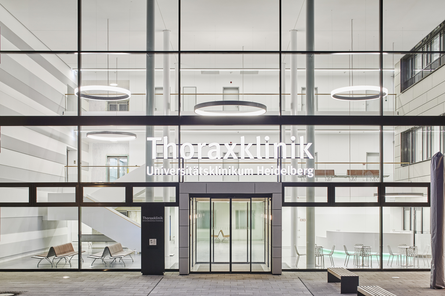 Eingangshalle & Treppenhaus Thoraxklinik mit CIRCOLO Ringleuchten Querbild: Außenansicht durch Glasfront auf Eingangshalle der Thoraxklinik mit Säulen und großen Ringleuchten abgehangen auf verschiedene Ebenen