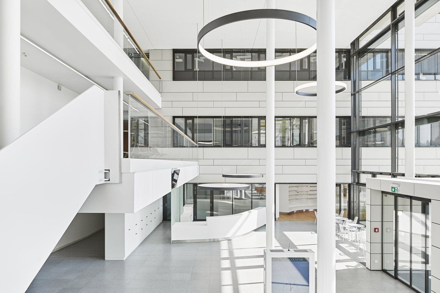 Empfangstheke mit Akustikleuchte CIRCOLO L+A - Eingangshalle & Treppenhaus Thoraxklinik mit CIRCOLO Ringleuchten Querbild: Hohe weiße Eingangshalle der Thoraxklinik mit Blick auf die Empfangstheke, über der zwei runde Leuchten mit Akustikfolie zum Sichtschutz hängen, im Raum Säulen und 2 große schwarze Ringleuchten nach innenstrahlend (CIRCOLO I) und 2 Ringleuchten (CIRCOLO INVERSE) nach außenstrahlend im Raum abgehangen.