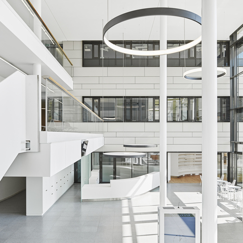 Reception desk with CIRCOLO L+A acoustic luminaires - Entrance hall & stairwell of thorax clinic with CIRCOLO ring luminaires Landscape: High white entrance hall of the Thorax Clinic with a view of the reception desk, above which two round luminaires with acoustic foil are suspended to provide privacy, in the room columns and 2 large black ring luminaires radiating inwards (CIRCOLO I) and 2 ring luminaires (CIRCOLO INVERSE) suspended in the room radiating outwards.