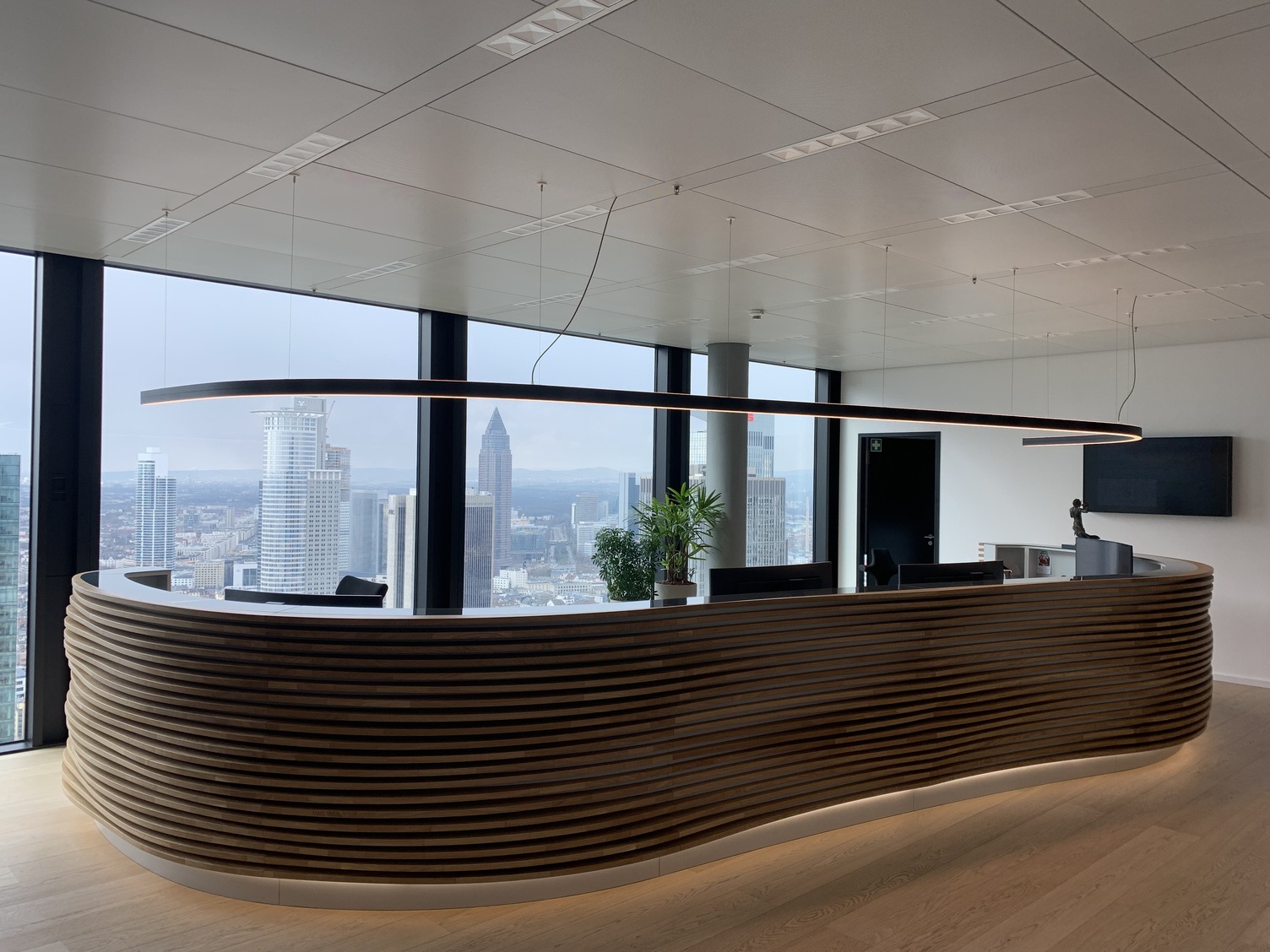 Maßgeschneiderte Pendelleuchte DUETTO über Empfangstresen Großer Empfangstresen vor einer großen Fensterfront im Bürogebäude von TRITON im Omni Tower Frankfurt. In der Form ist die lange Empfangstheke geschwungen und scheint optisch von außen mehrrippig aus dunklen Holz aufgebaut, darüber ist eine lineare schwarze Pendelleuchte DUETTO in gleicher geschwungener Form abgehangen. Maßgeschneiderte Sonderleuchte für die Kundentheke.