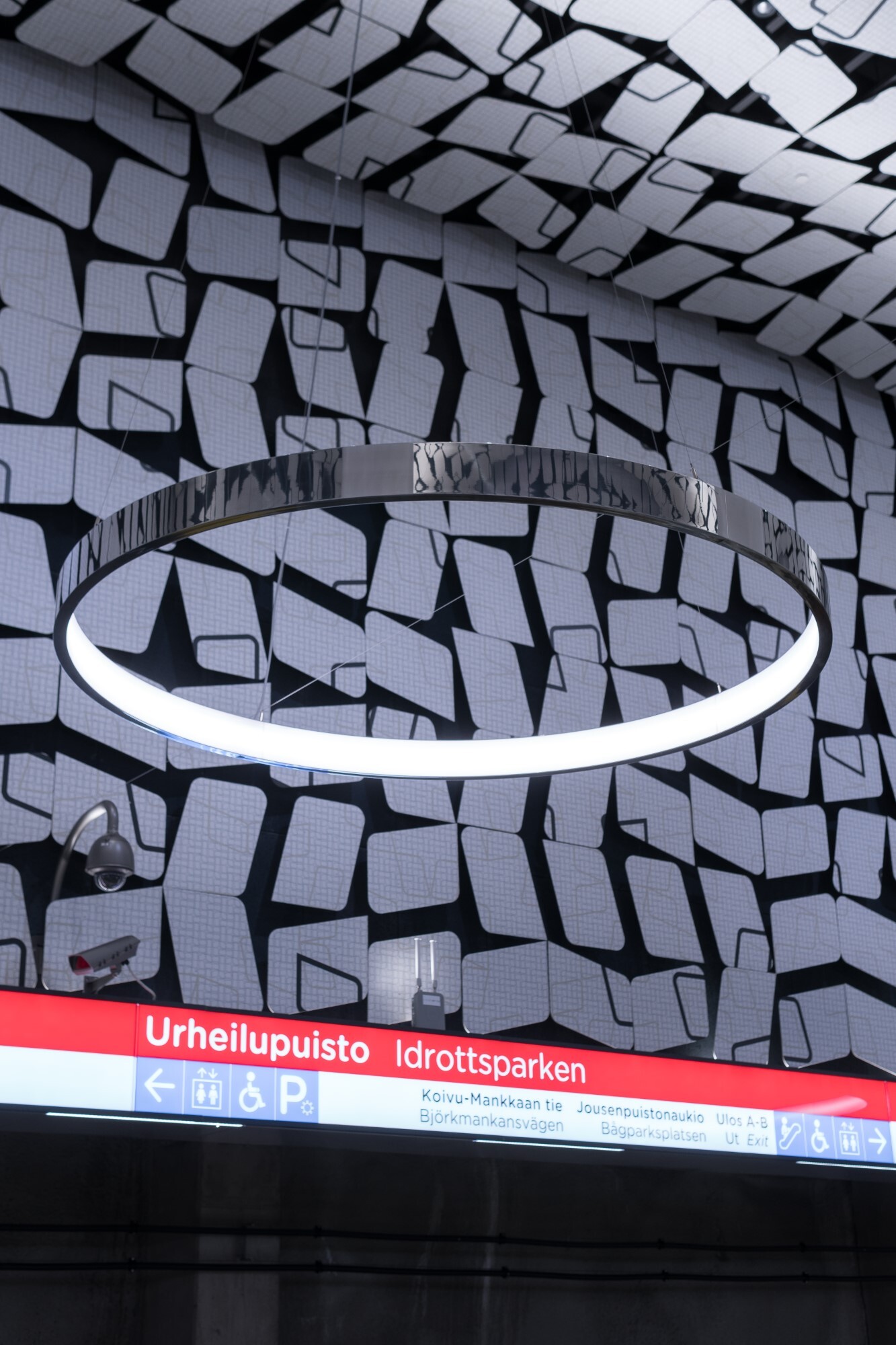 A black-and-white wall and ceiling with a polished aluminium ring lamp (CIRCOLO) in front of it, which reflects its surroundings.