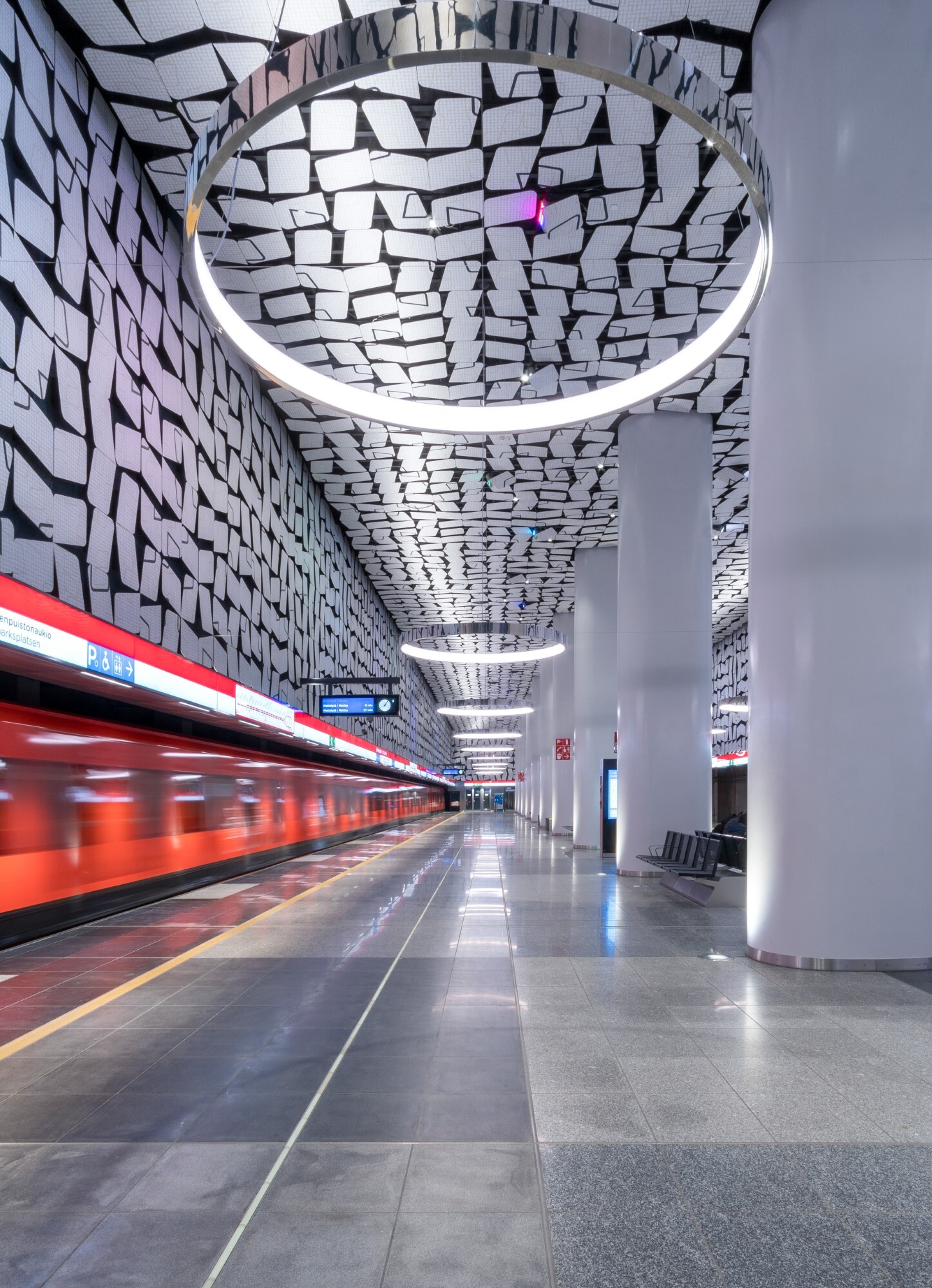 Orange underground train passing through the image, the underground station consists of graphic patterns on the walls and ceilings, which are reflected in the round CIRCOLO ring lights suspended from the ceiling.