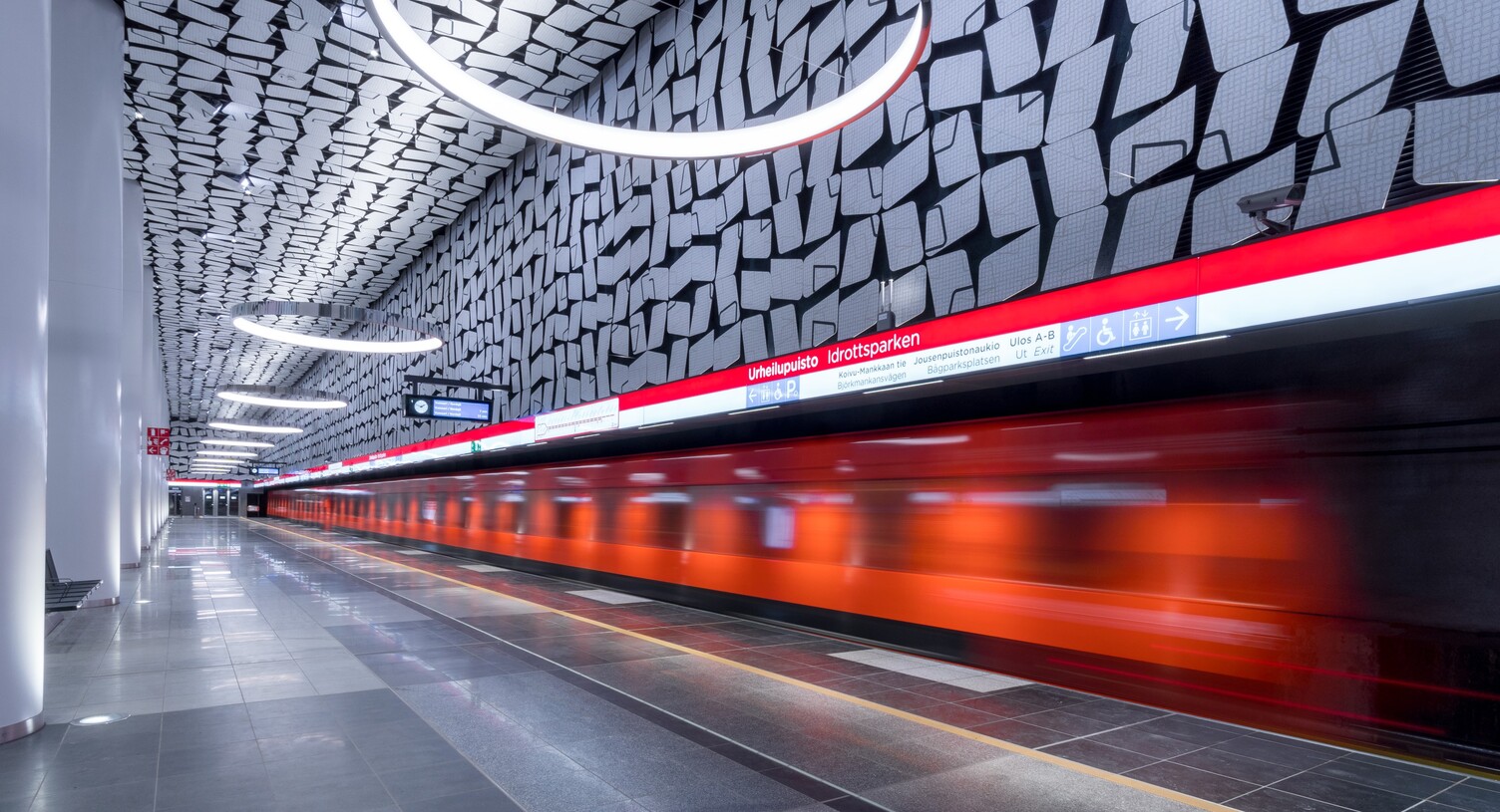 Orange underground train passing through the image, the underground station consists of graphic patterns on the walls and ceilings, which are reflected in the round CIRCOLO ring lights suspended from the ceiling.