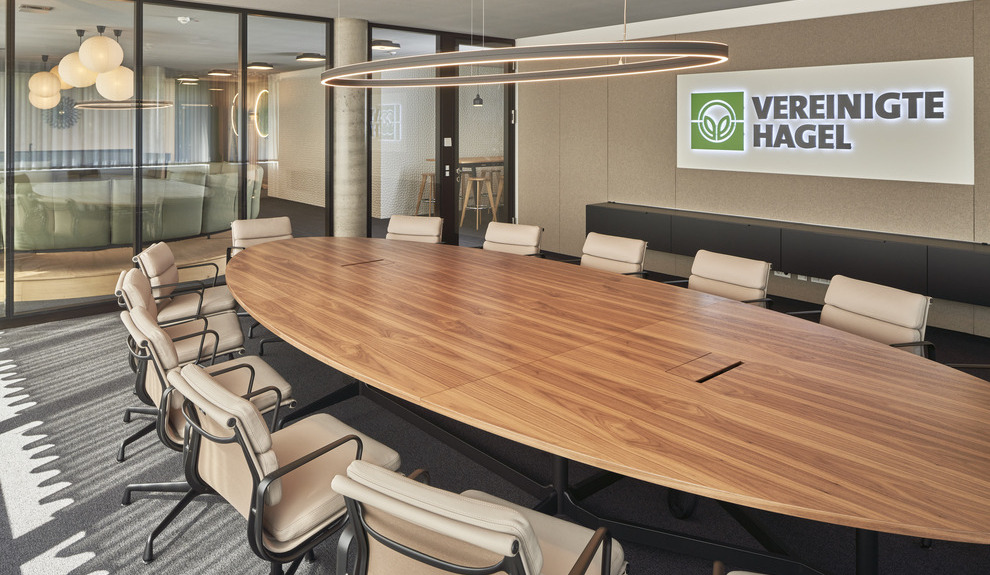 Meeting room at Vereinigte Hagel with DOPPIO ELLIPSE light Meeting room with DOPPIO ELLIPSE light, oval wooden table, white chairs, and company logo on the wall.