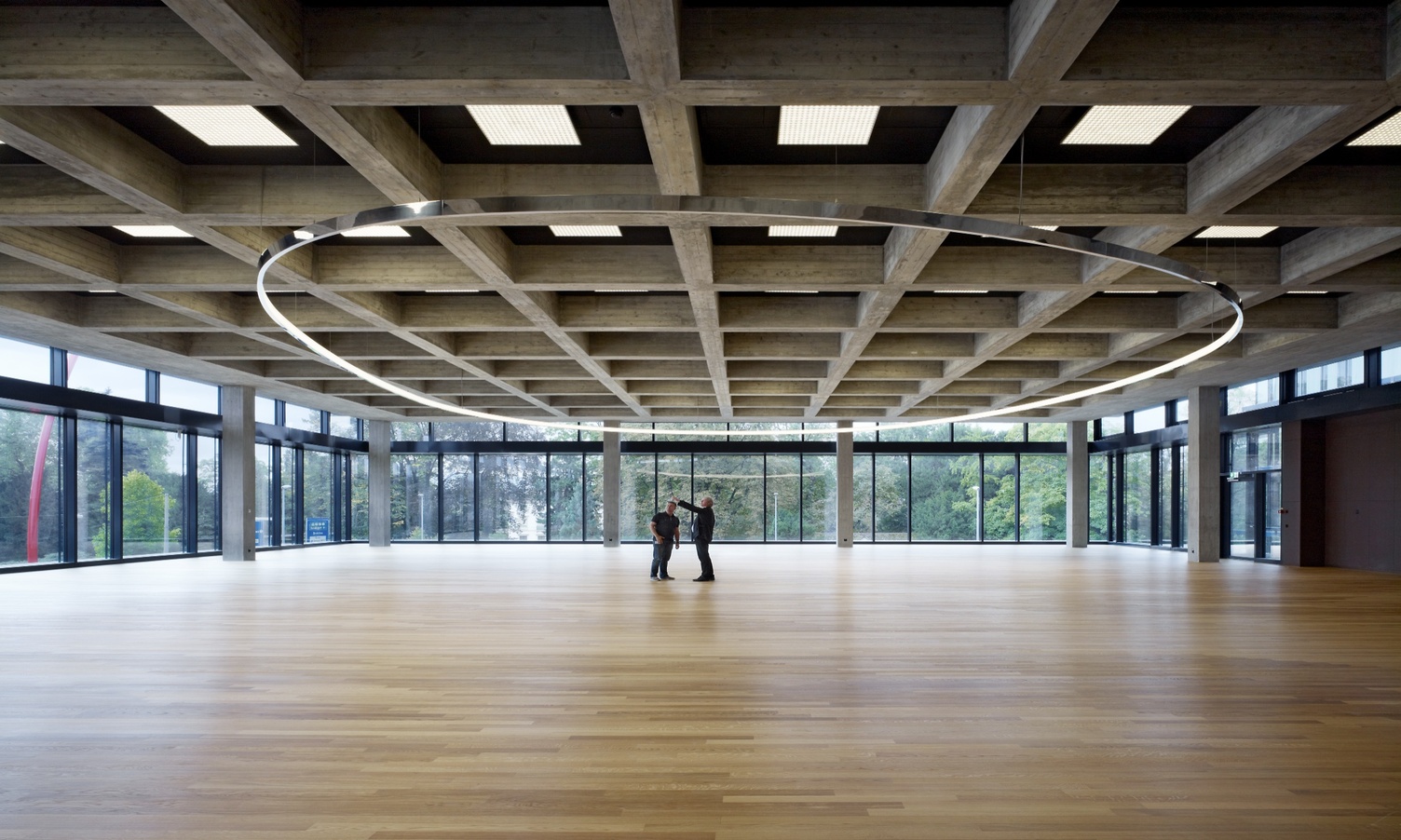 Elliptische Pendel-Ring-Sonderleuchte von SATTLER bei Roche Elliptische Pendel-Ring-Sonderleuchte (19m) Freiform in der noch großen leeren Kantine der Roche AG, Bau 67, Basel mit zwei Personen darunter stehend.
