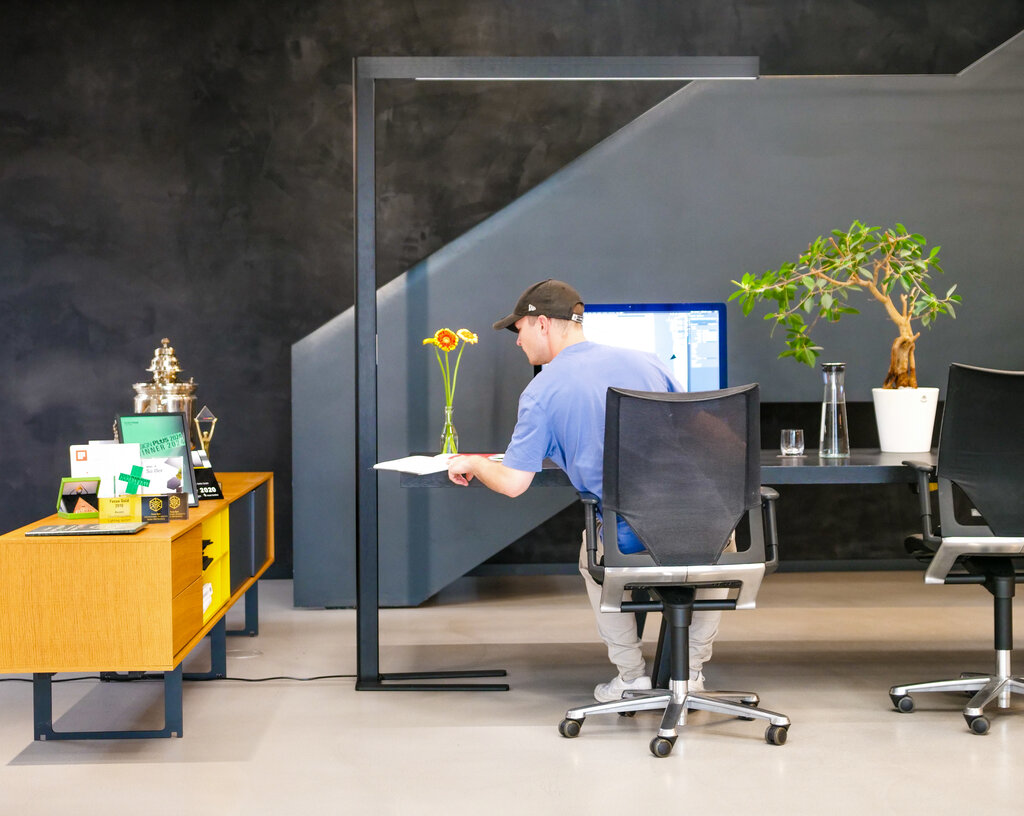 A large desk with a linear floor lamp. A young man with a cap sits on the office chair and realises his creative ideas on the screen.