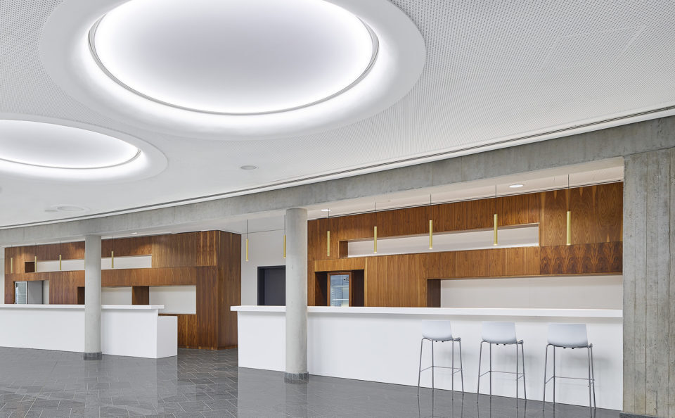 Lounge area in gold colours with benches as alcoves in a large hall with a white ceiling in which large round light fields are arranged. Large hall with a stone floor and a white ceiling in which large round light fields are arranged, the view goes into a bar area, which is designed as niches and is emphasised by brown wooden walls, in addition, golden bar lights (TIAs) are suspended at regular intervals over the entire bar.